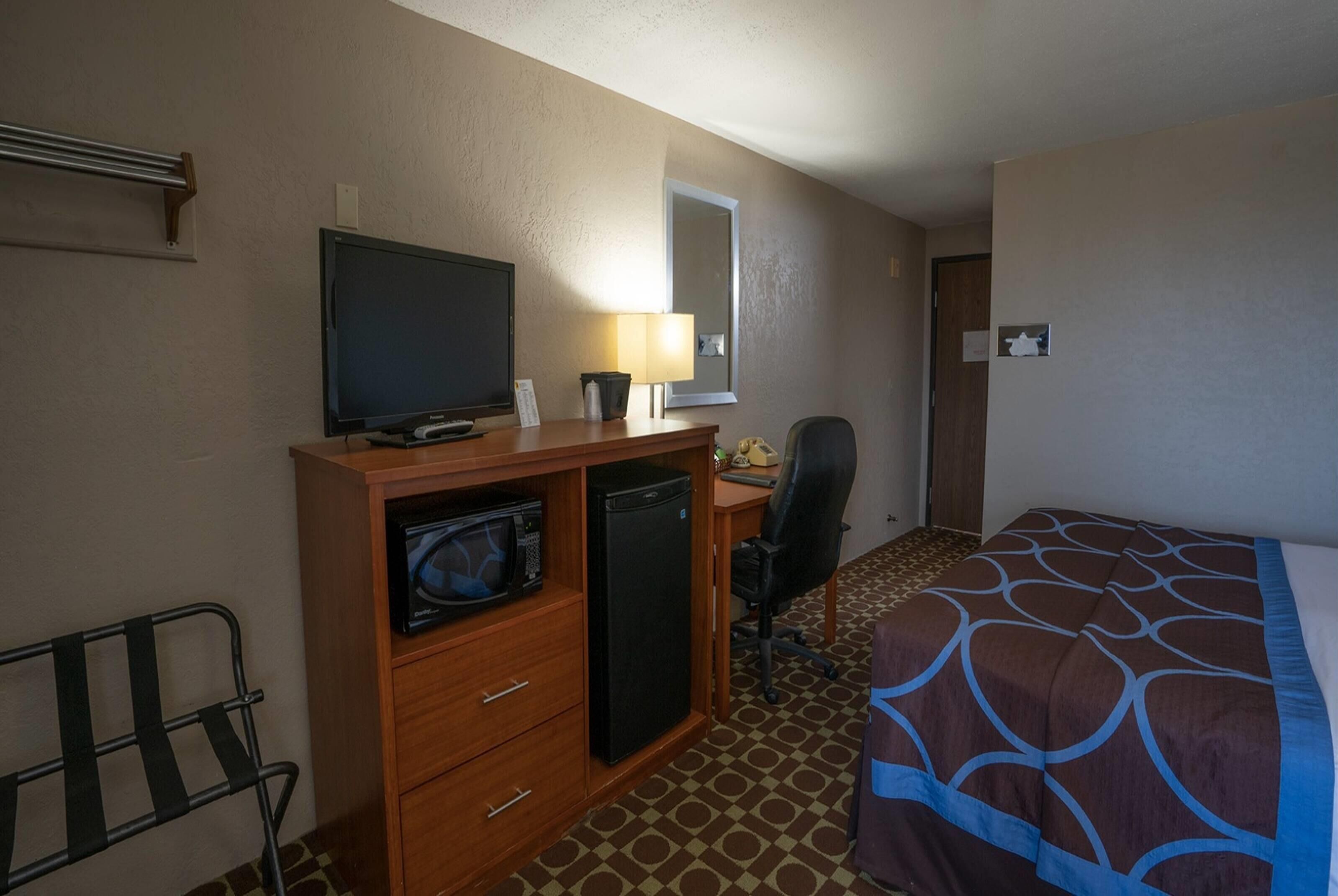 Habitación de hotel con una cama, un escritorio con computadora, televisor, microondas, una silla y un refrigerador pequeño.