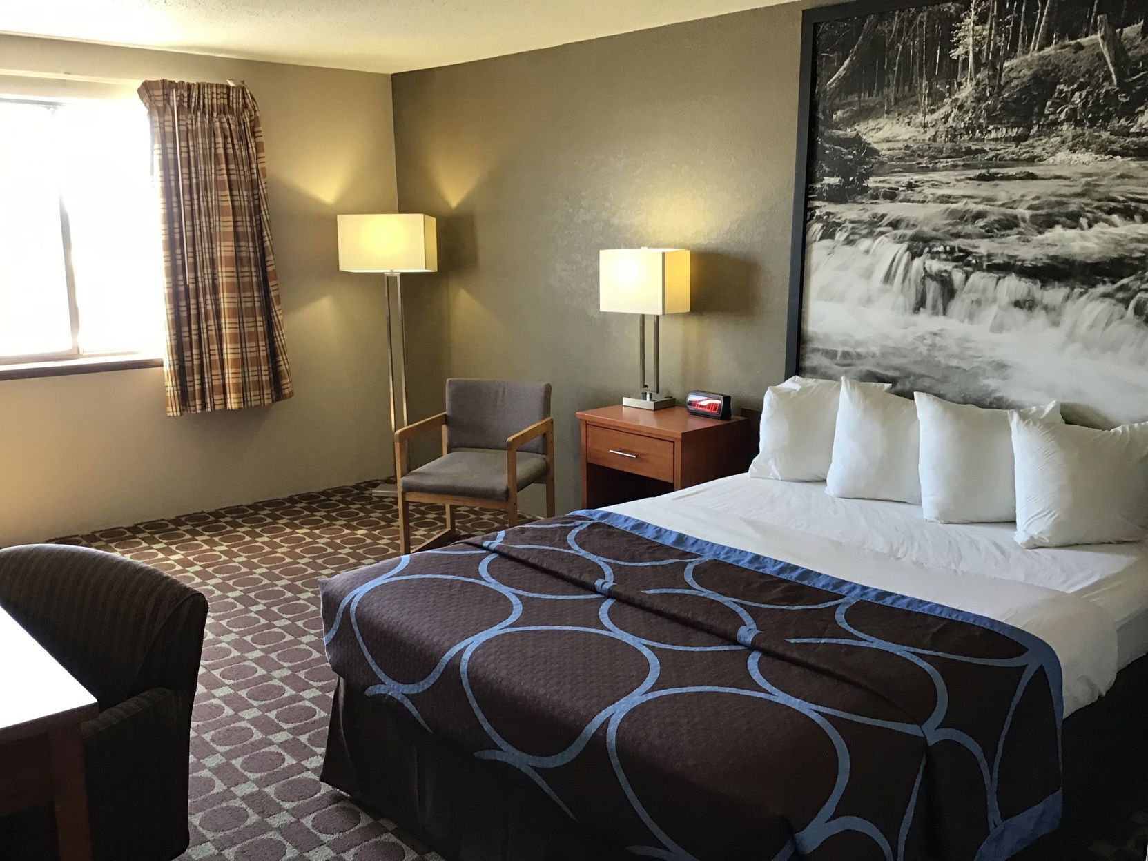 A hotel room with a bed, two bedside lamps, a chair, a desk, and a large painting of a waterfall.