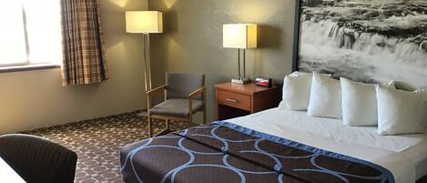 Ein Hotelzimmer mit einem Bett, zwei Nachttischen, einem Sessel, einem Schreibtisch, einer Lampe und einem großen Gemälde eines Wasserfalls.