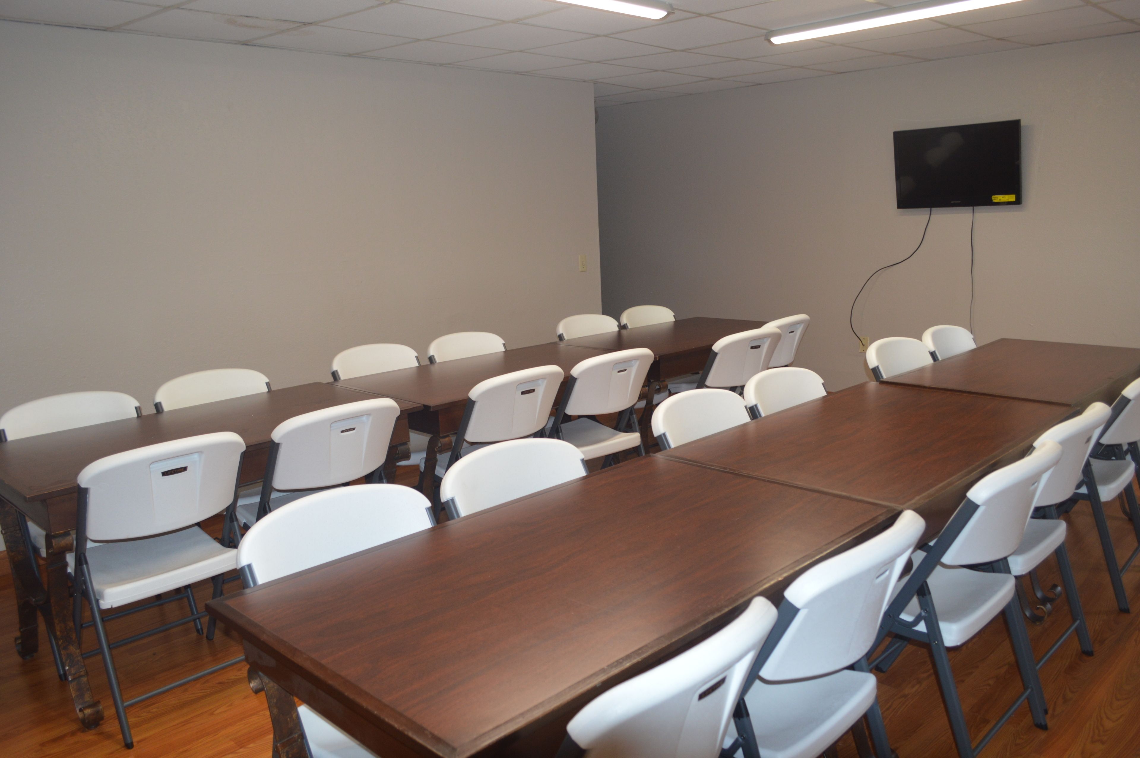 A conference room with a long table and white chairs, a flat-screen TV mounted on the wall, and a ceiling light fixture.