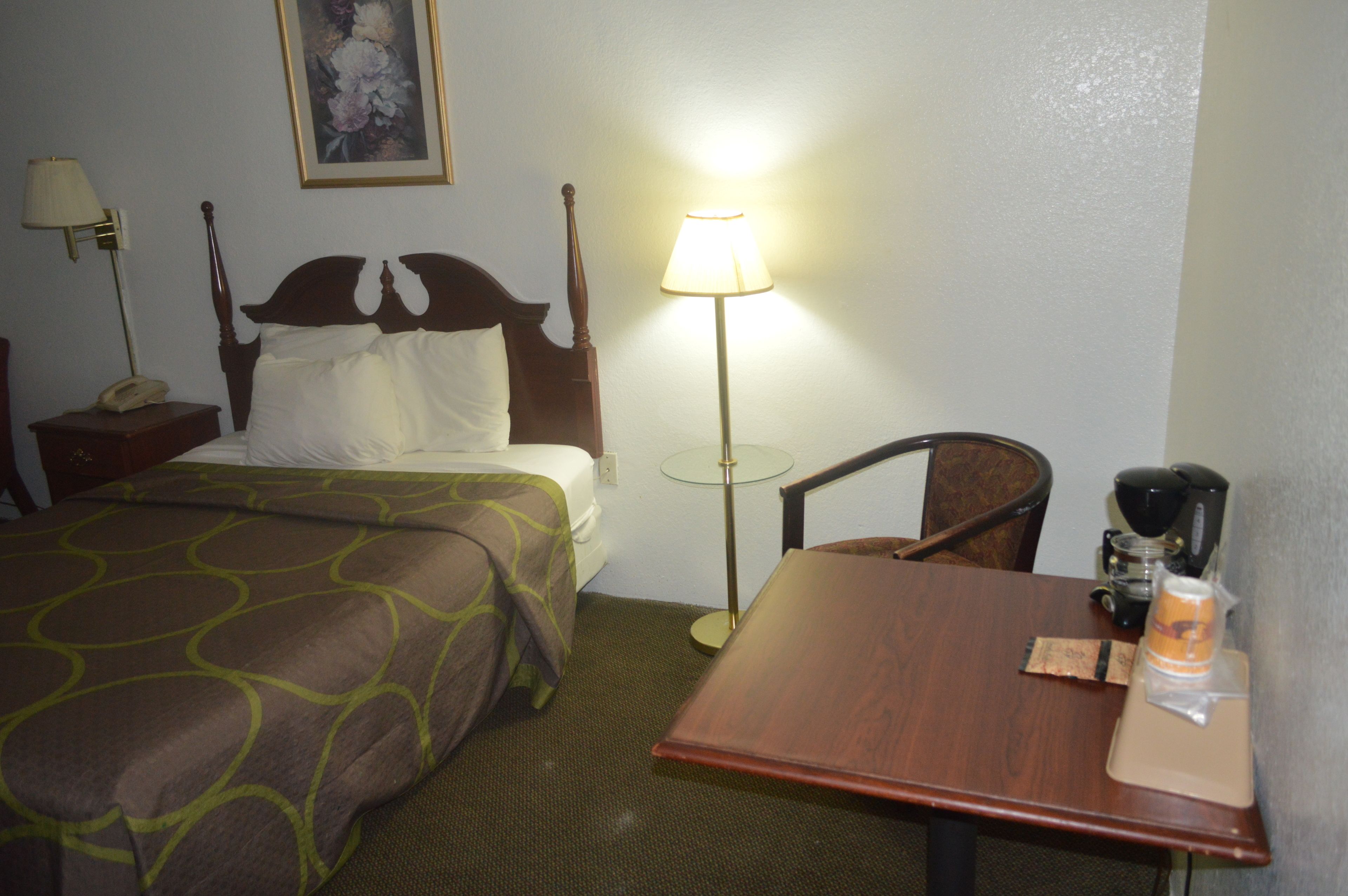 Ein Hotelzimmer mit Bett, Nachttisch, Lampe, Sessel und Tisch mit Kaffeemaschine und Getränkebehälter.