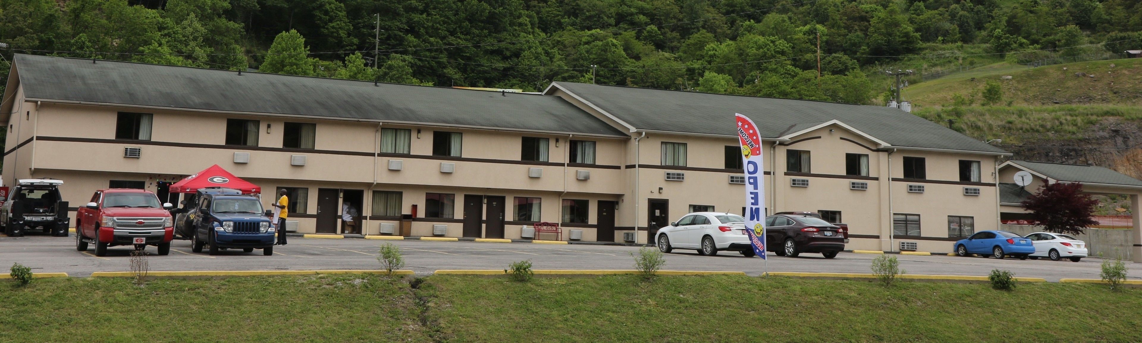 Un motel con una fila de habitaciones, un estacionamiento y una colina al fondo.