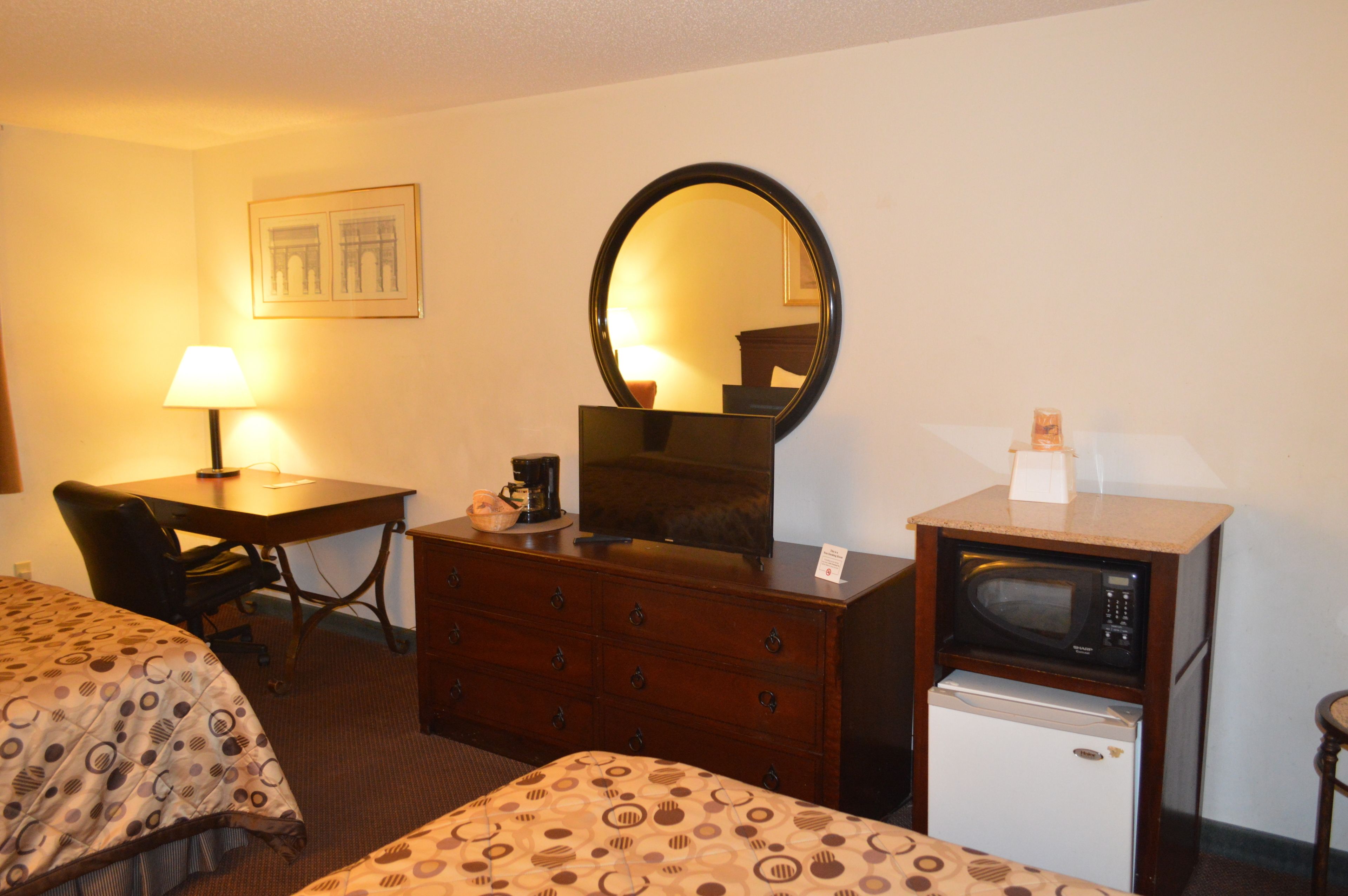 A hotel room with two beds, a desk with a lamp, a dresser, a mirror, a microwave, and a refrigerator.