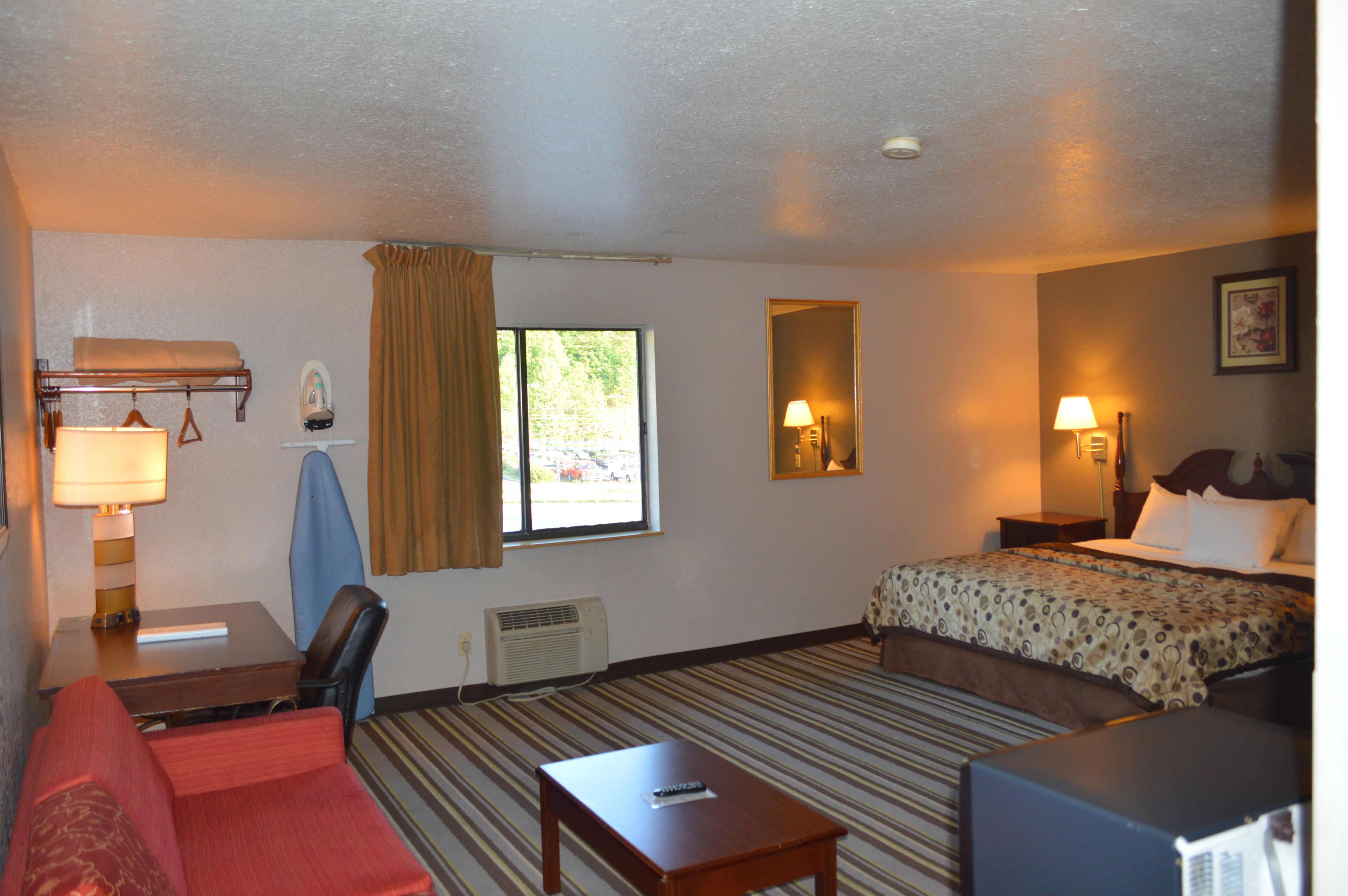 A hotel room with a bed, a desk with a lamp, a chair, a small table, and a window with curtains.