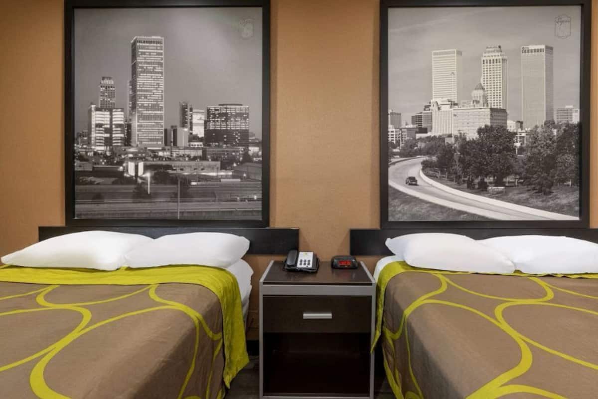 Habitación de hotel con dos camas, cada una con un almohadón blanco y una colcha marrón con detalles amarillos. Hay una mesita de noche entre las camas con un teléfono y una pequeña mesa. Dos fotografías en blanco y negro de un paisaje urbano se encuentran en la pared sobre las camas.