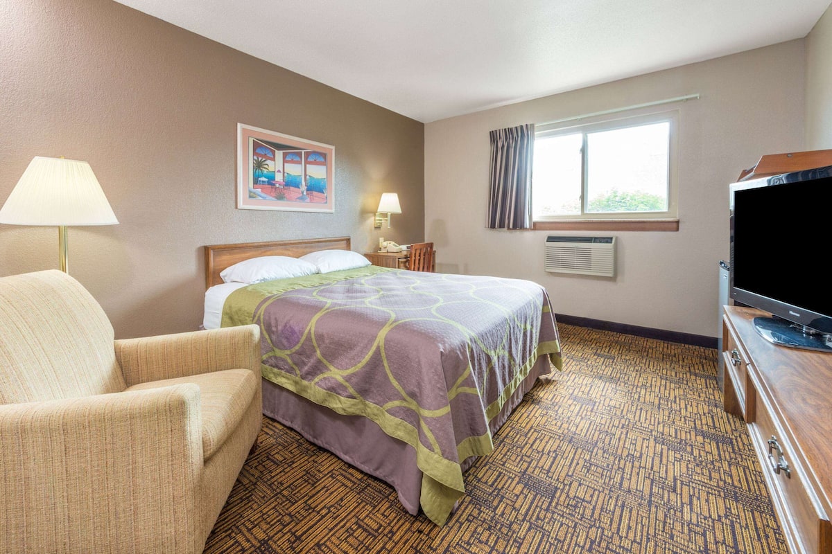 A hotel room with a bed, a chair, a television, a desk with a lamp, and a window with curtains.