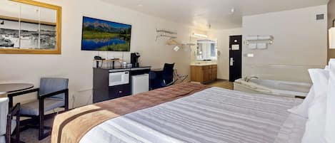 A hotel room with a large bed, a desk with a chair, a small kitchenette, a bathroom with a bathtub, and a large window with a view of mountains.