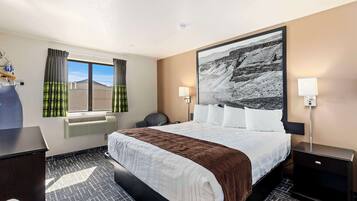 Una camera d'albergo con un letto grande, una scrivania, una sedia, una finestra con tende, un condizionatore d'aria a muro e un quadro incorniciato di grandi dimensioni.