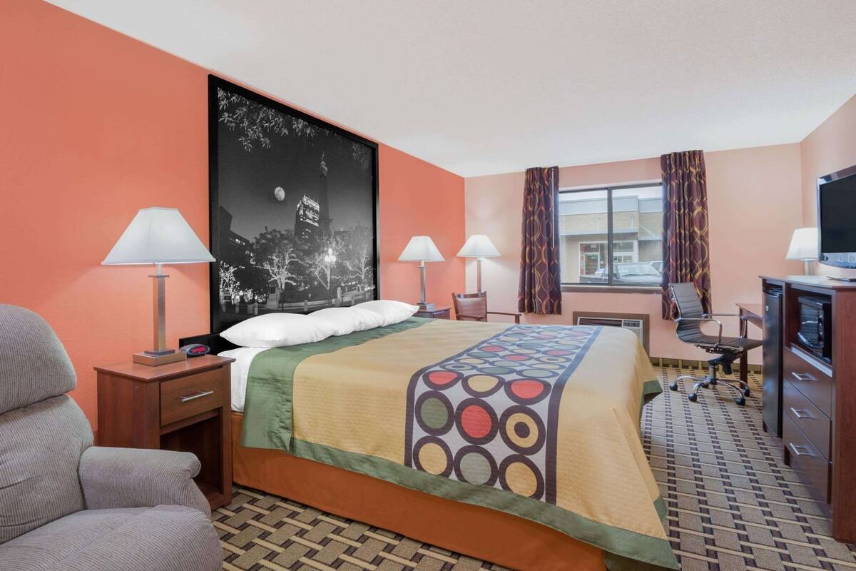 Een hotelkamer met een bed, twee nachtkastjes met lampen, een bureau met stoel, een televisie en een groot raam met gordijnen.