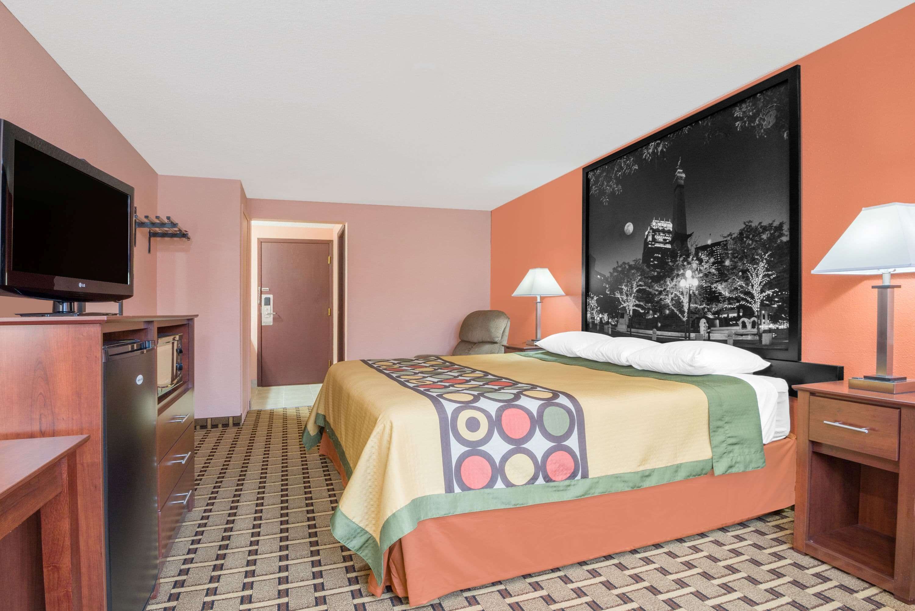 Una habitación de hotel con una cama, televisión, escritorio, silla y mesita de noche con lámpara.