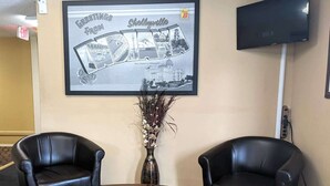 Un vestíbulo con dos sillones de cuero negro, una mesa de madera, un cartel de bienvenida enmarcado, un jarrón con flores secas y un televisor montado en la pared.