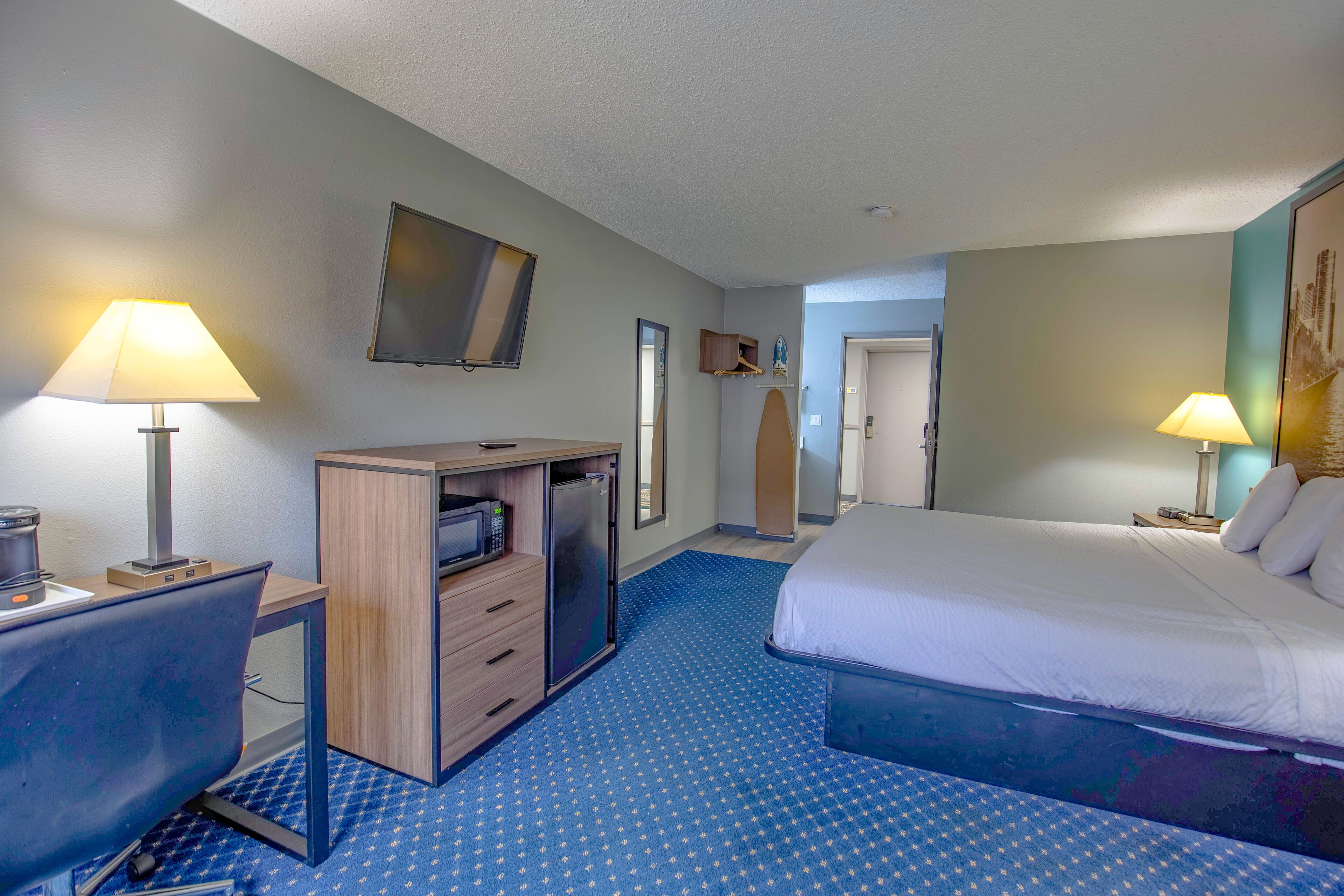 Quarto de hotel com cama, escrivaninha e cómoda. Há uma televisão na parede e um quadro sobre a cama.