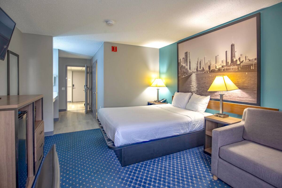 Habitación de hotel con una cama grande, dos lámparas de noche, una silla, un escritorio y un cuadro con una vista urbana.