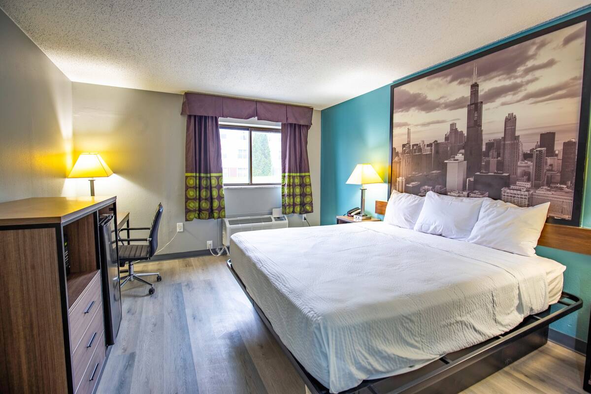 Habitación de hotel con cama, escritorio, silla y una gran pintura de un paisaje urbano en la pared.