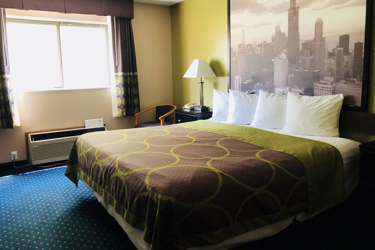 Una habitación de hotel con cama, una silla, un escritorio, una lámpara, ventana con cortinas y un cartel con un paisaje urbano en la pared.