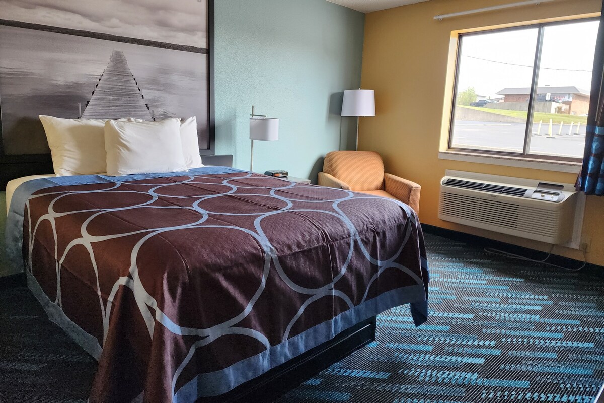A hotel room with a bed, a chair, a window, and a wall-mounted air conditioner.