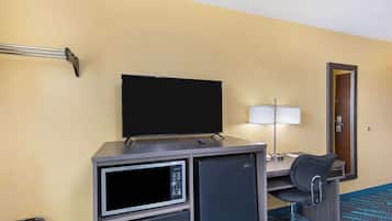 Habitación de hotel con televisión de pantalla plana, mini refrigerador, microondas, escritorio con lámpara, una silla y una puerta.