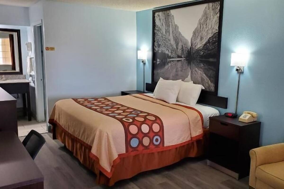 Habitación de hotel con cama, escritorio, silla, mesita de noche con teléfono y un cuadro grande en la pared.