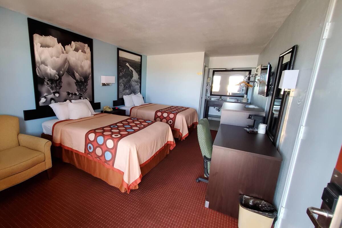 Habitación de hotel con dos camas, un escritorio, una silla, un cesto de basura y un baño visible al fondo.
