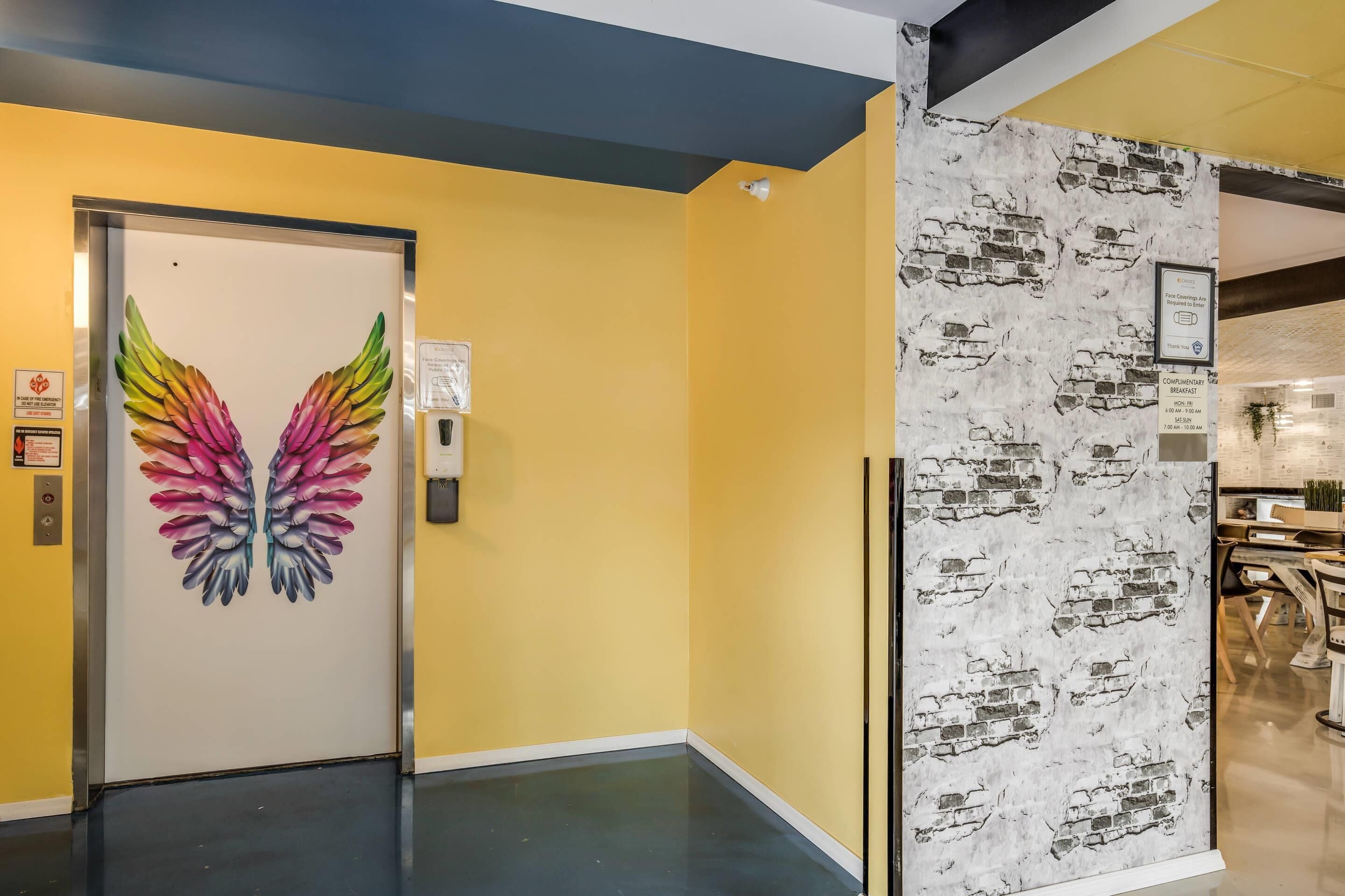 A hallway with a door featuring a colorful feather design, a wall with a brick pattern, and a sign indicating complimentary breakfast hours.