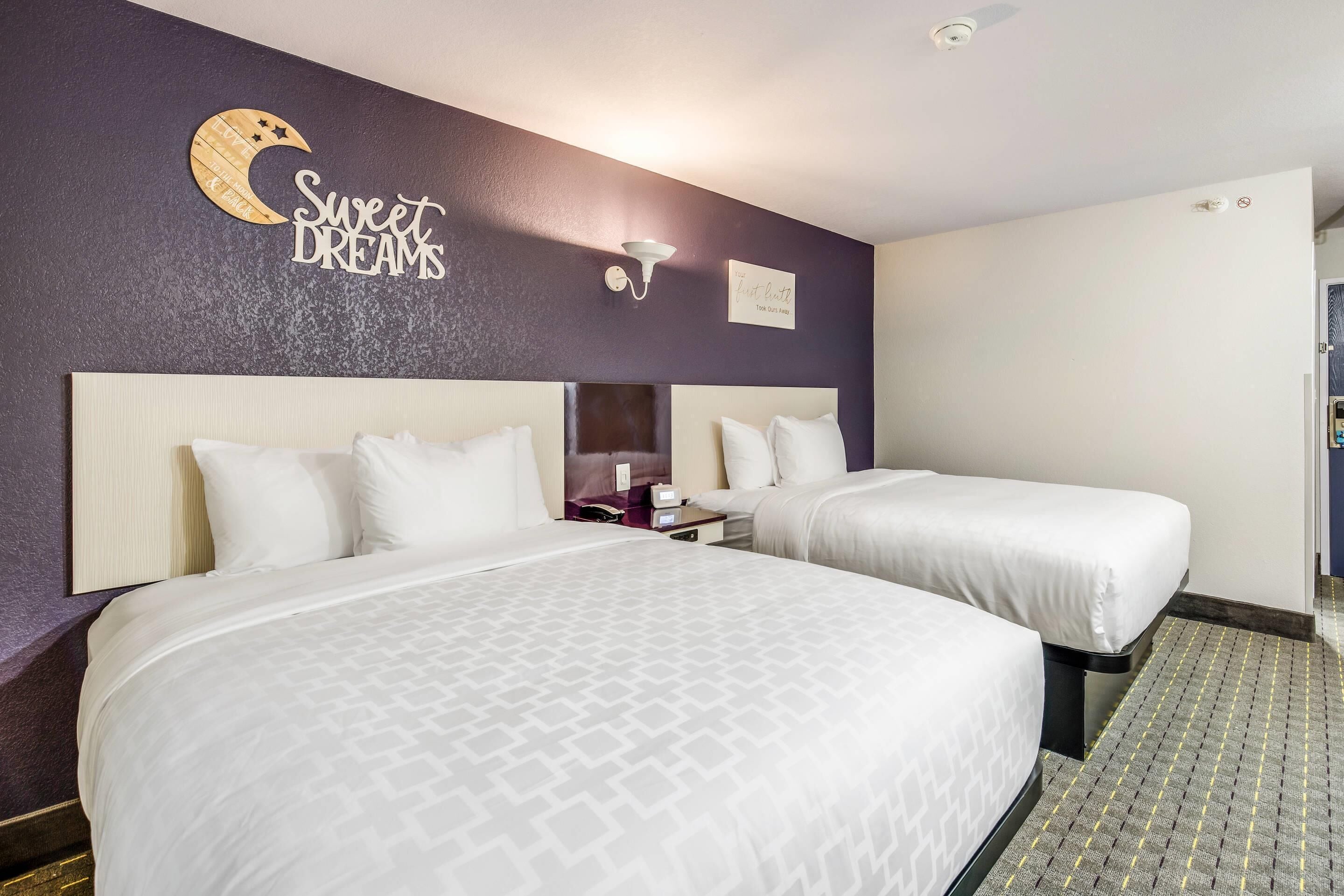 A hotel room with two beds, a nightstand, a wall clock, and a sign that reads 'Sweet Dreams'.