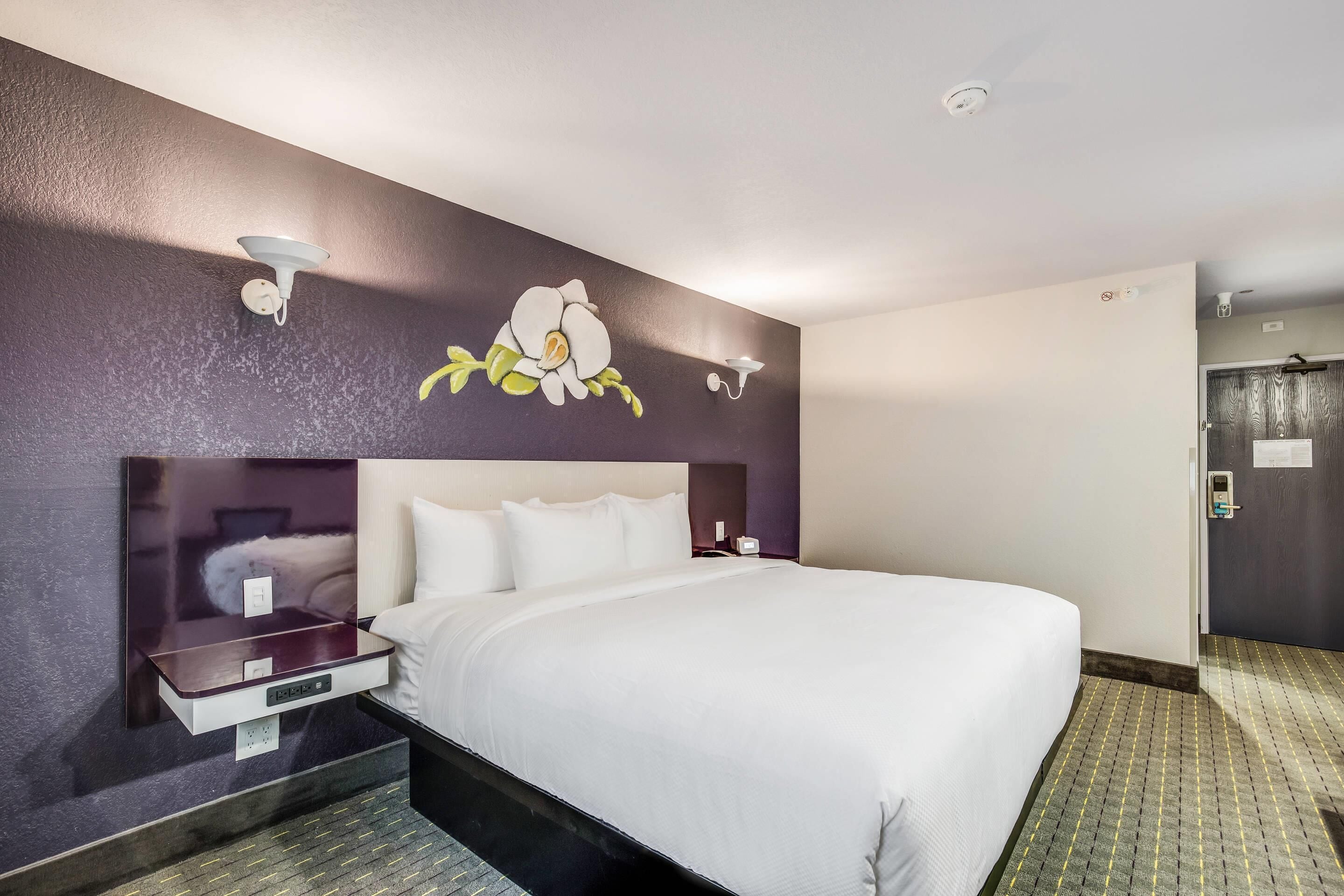 A hotel room with a large bed, a nightstand, a wall-mounted lamp, and a door with a lock.