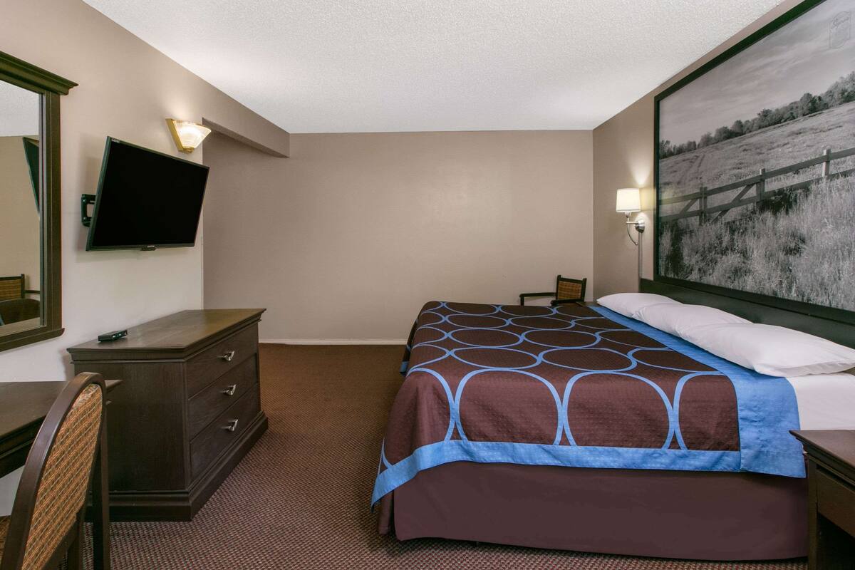A hotel room with a bed, a dresser, a television mounted on the wall, and a painting on the wall.