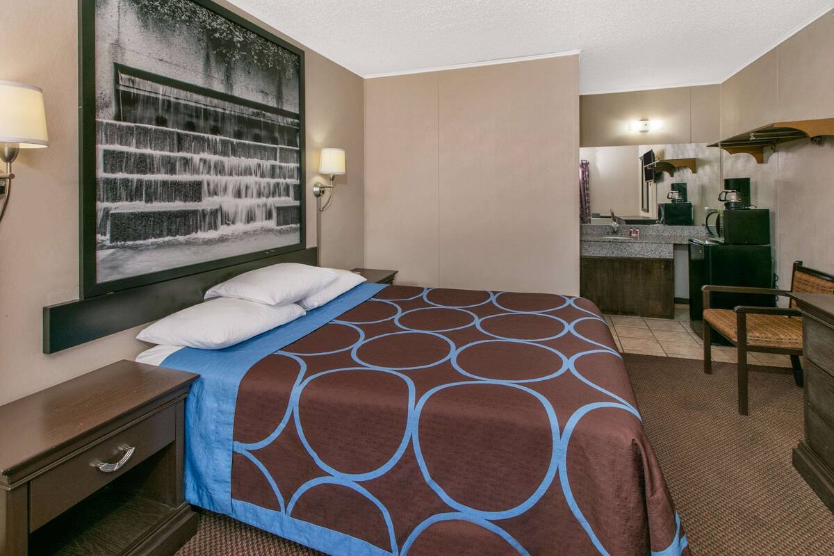 A hotel room with a bed, a desk, a chair, a large painting, a kitchenette, and a bathroom.