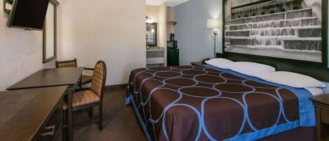 Quarto de hotel com cama, escrivaninha, cadeira, televisão, quadro e mesa de cabeceira.