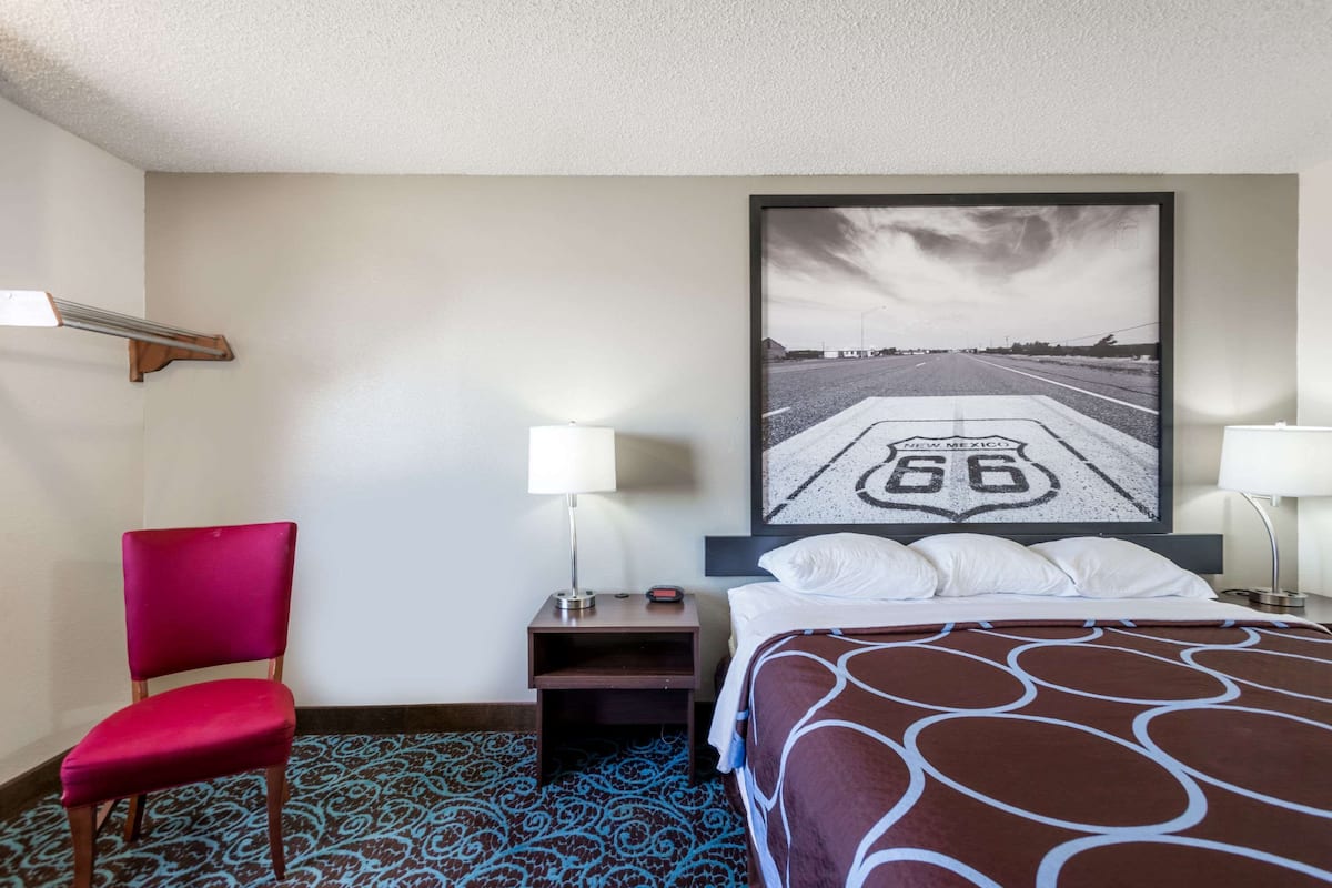 Ein Hotelzimmer mit Bett, Stuhl, Nachttisch, zwei Lampen, einem an der Wand hängenden Bild einer Straße und einem gemusterten Teppich.