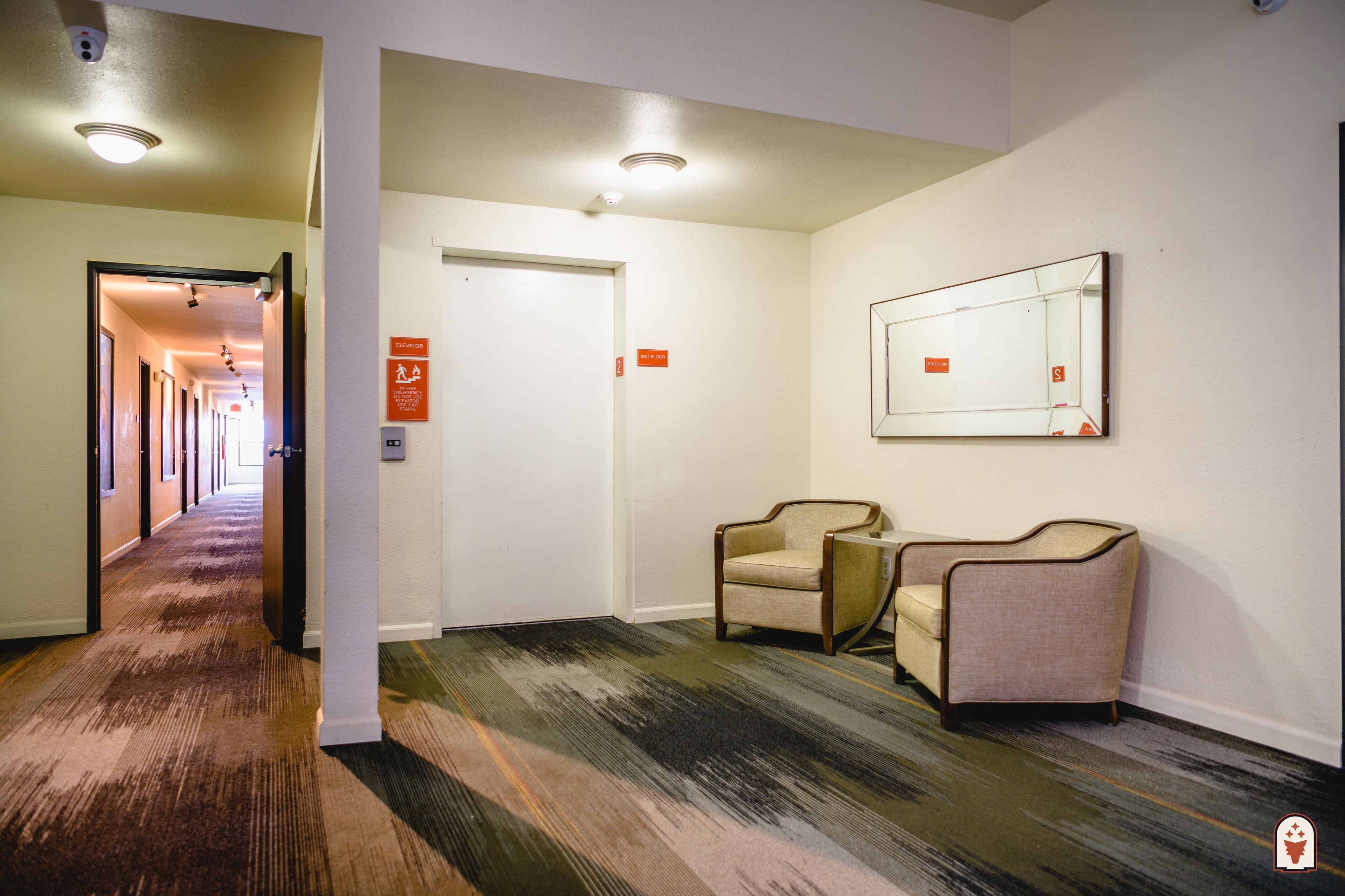 A hotel lobby with a carpeted floor, two armchairs, a mirror on the wall, and a door with an elevator sign.