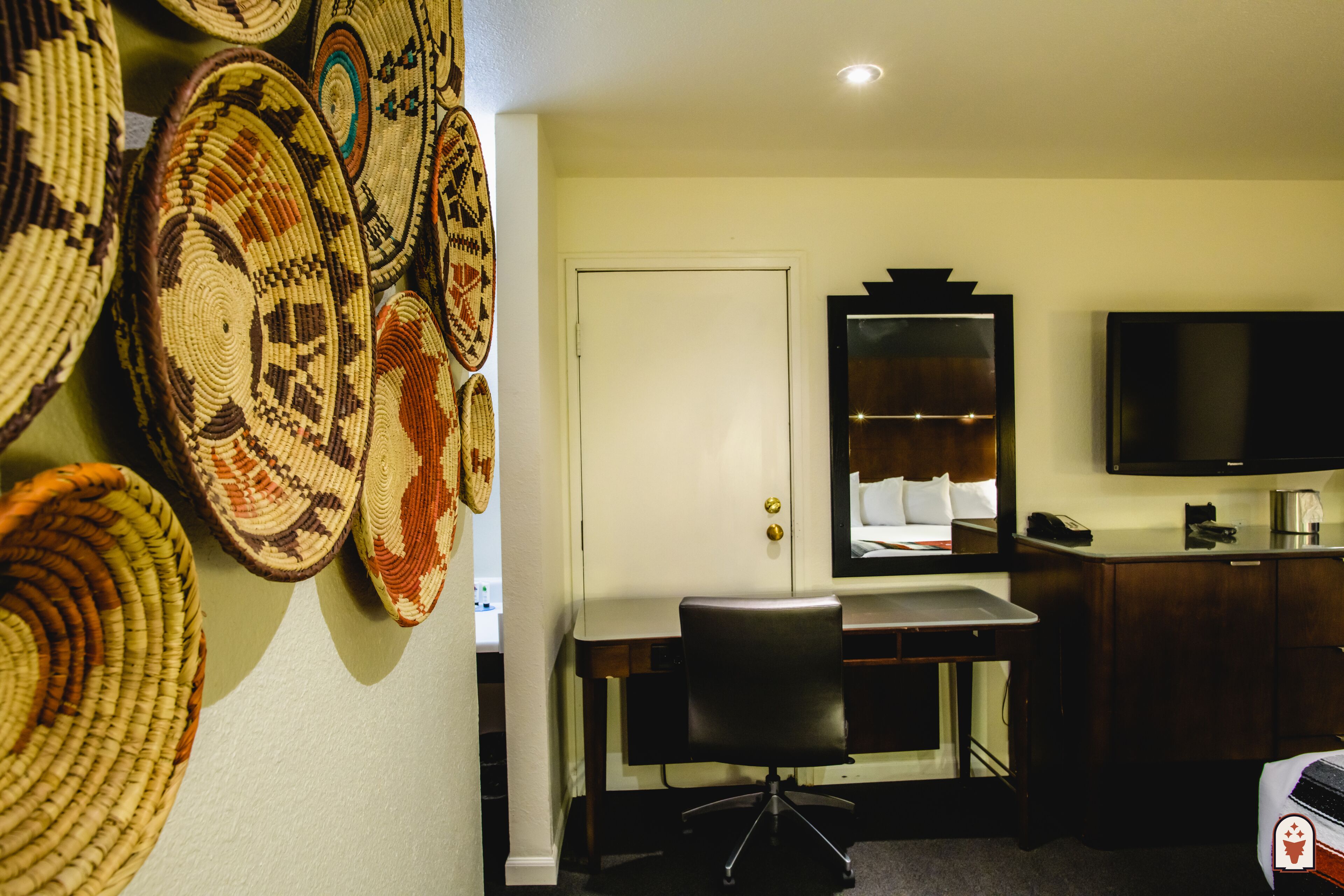 A hotel room with a bed, a desk with a chair, a television, and a wall adorned with woven baskets.