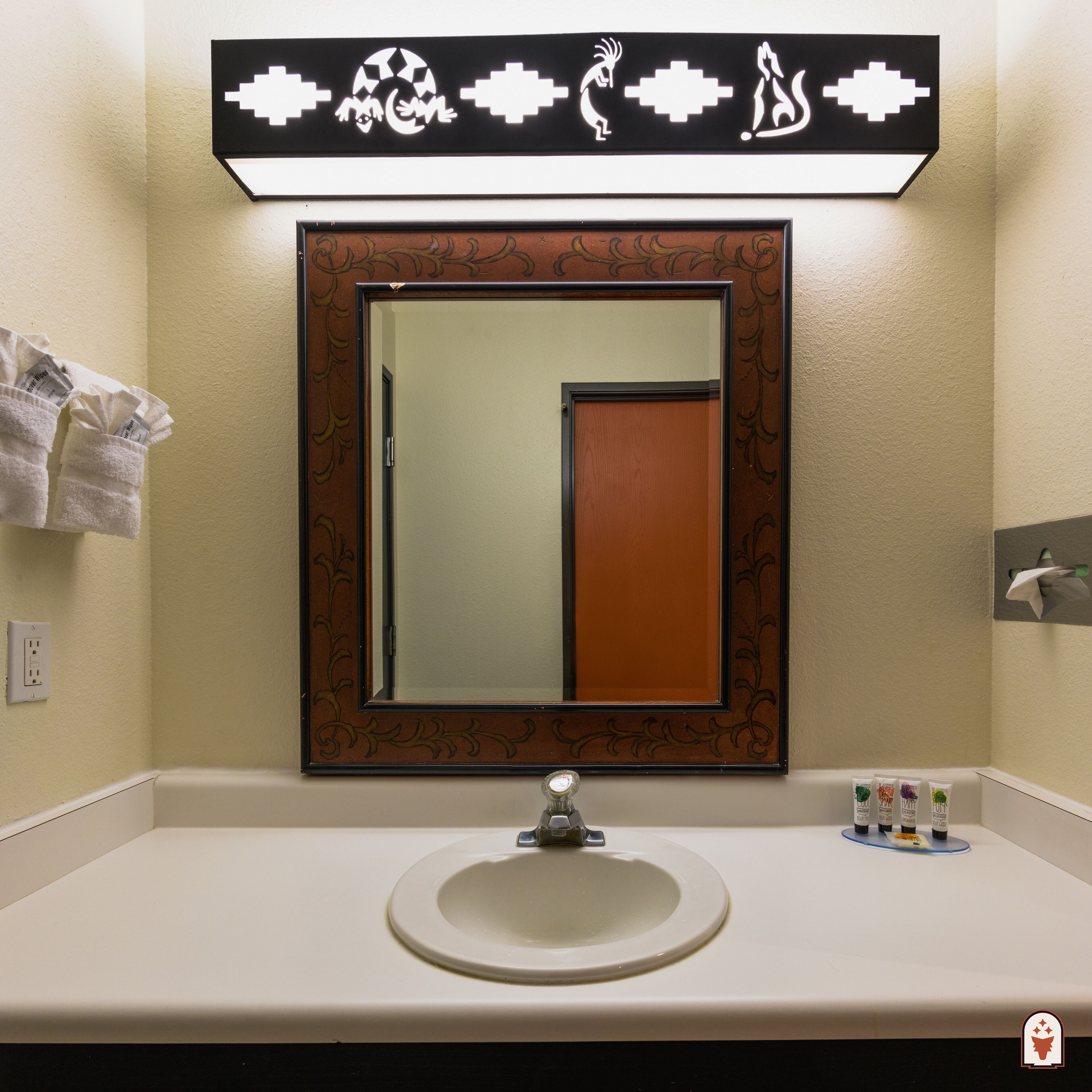 A bathroom with a round sink, a mirror with a decorative frame, and a wall-mounted light fixture above the mirror.
