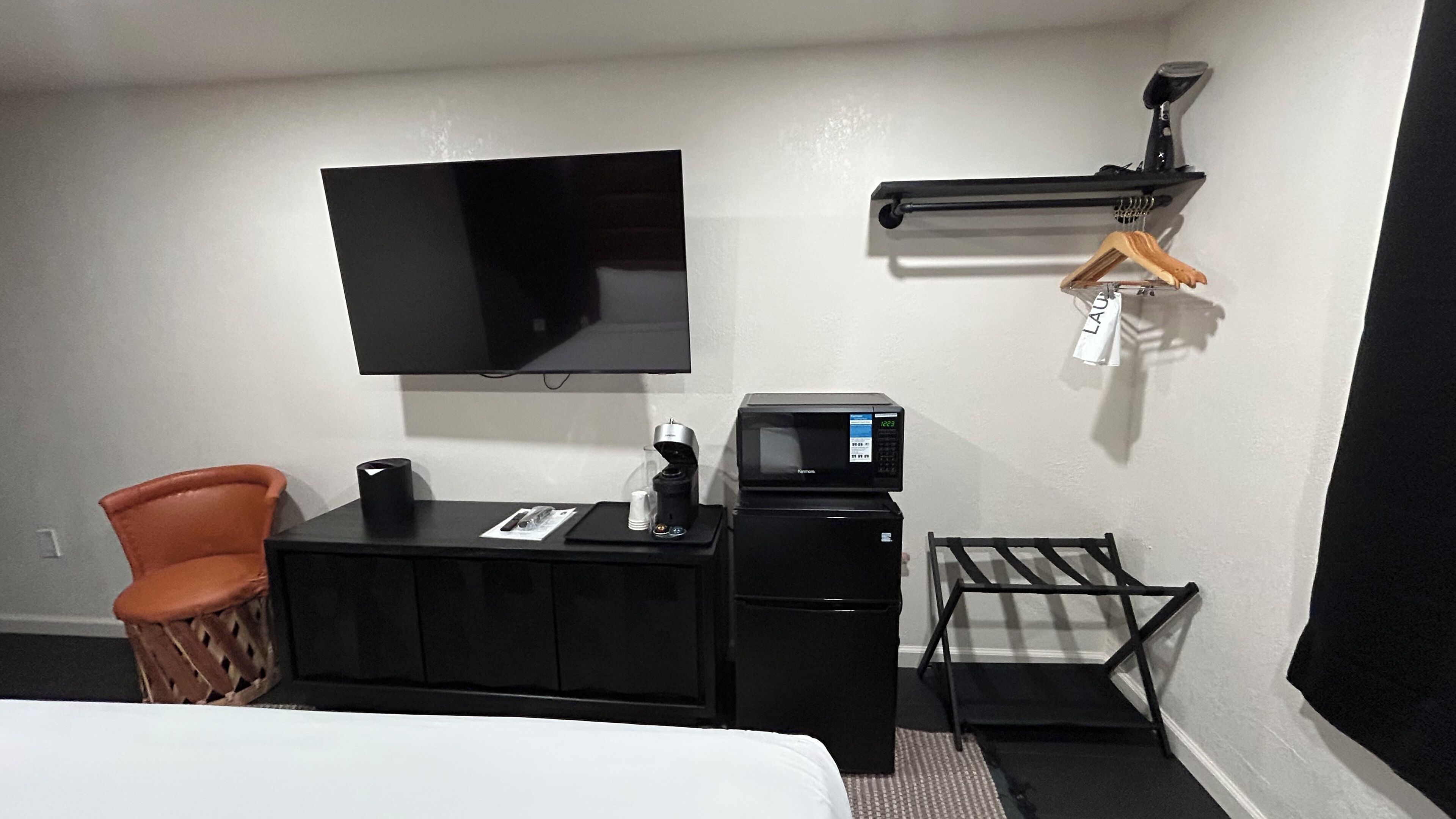 A hotel room with a bed, a desk with a coffee machine, a TV, a small refrigerator, and a clothes drying rack.
