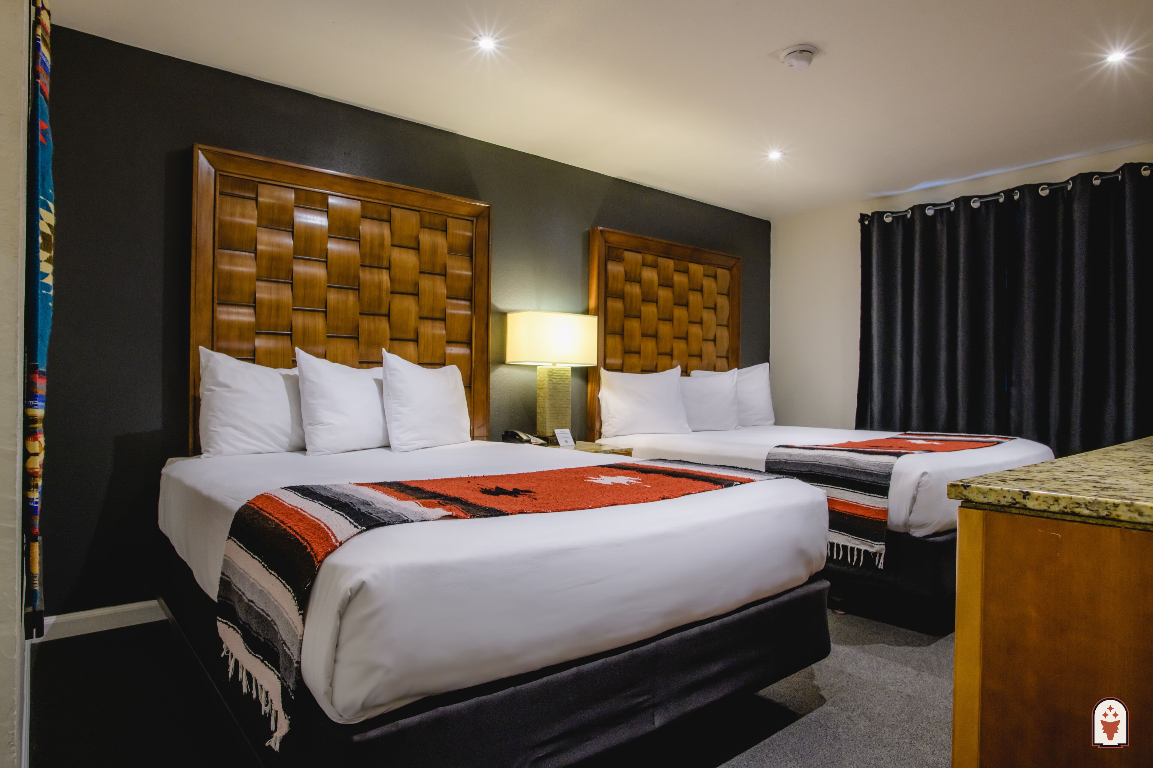 A hotel room with two beds, each with a wooden headboard, white bedding, and a red and black striped blanket. There is a nightstand with a lamp between the beds, and a black curtain on the window.