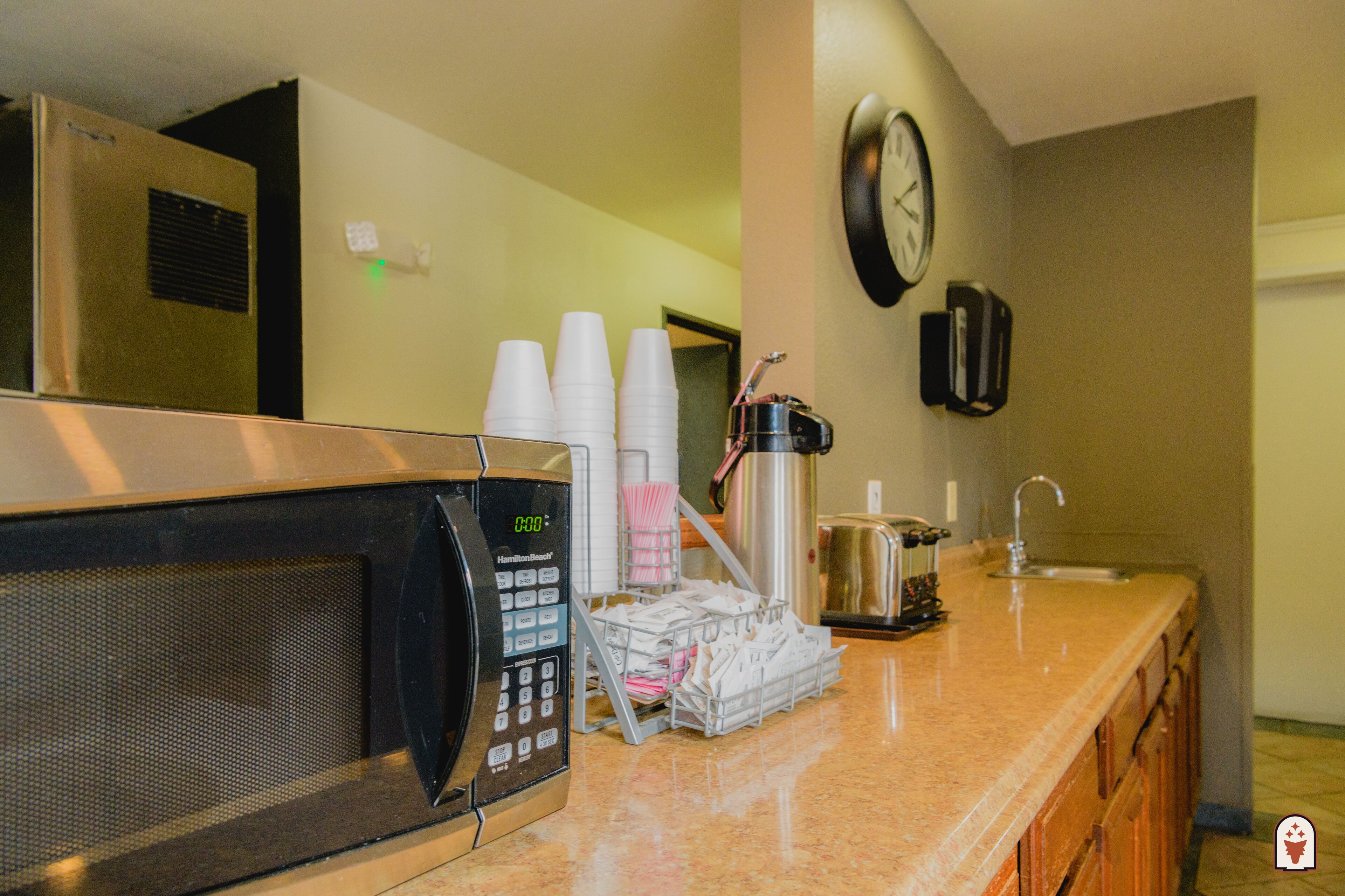 A kitchen with a microwave, coffee cups, a coffee maker, a kettle, and a clock on the wall.