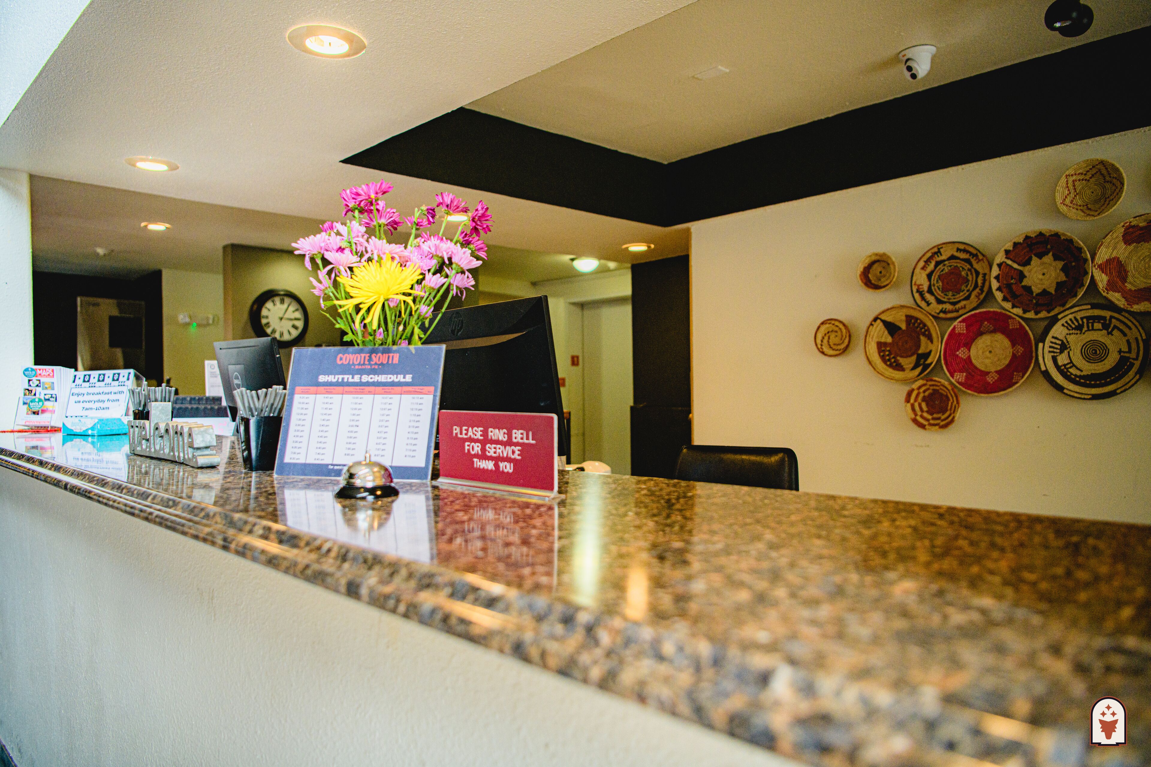 Un comptoir d’accueil d’hôtel avec une horloge, un vase de fleurs, un panneau indiquant les horaires de la navette et un mur décoré d’assiettes.