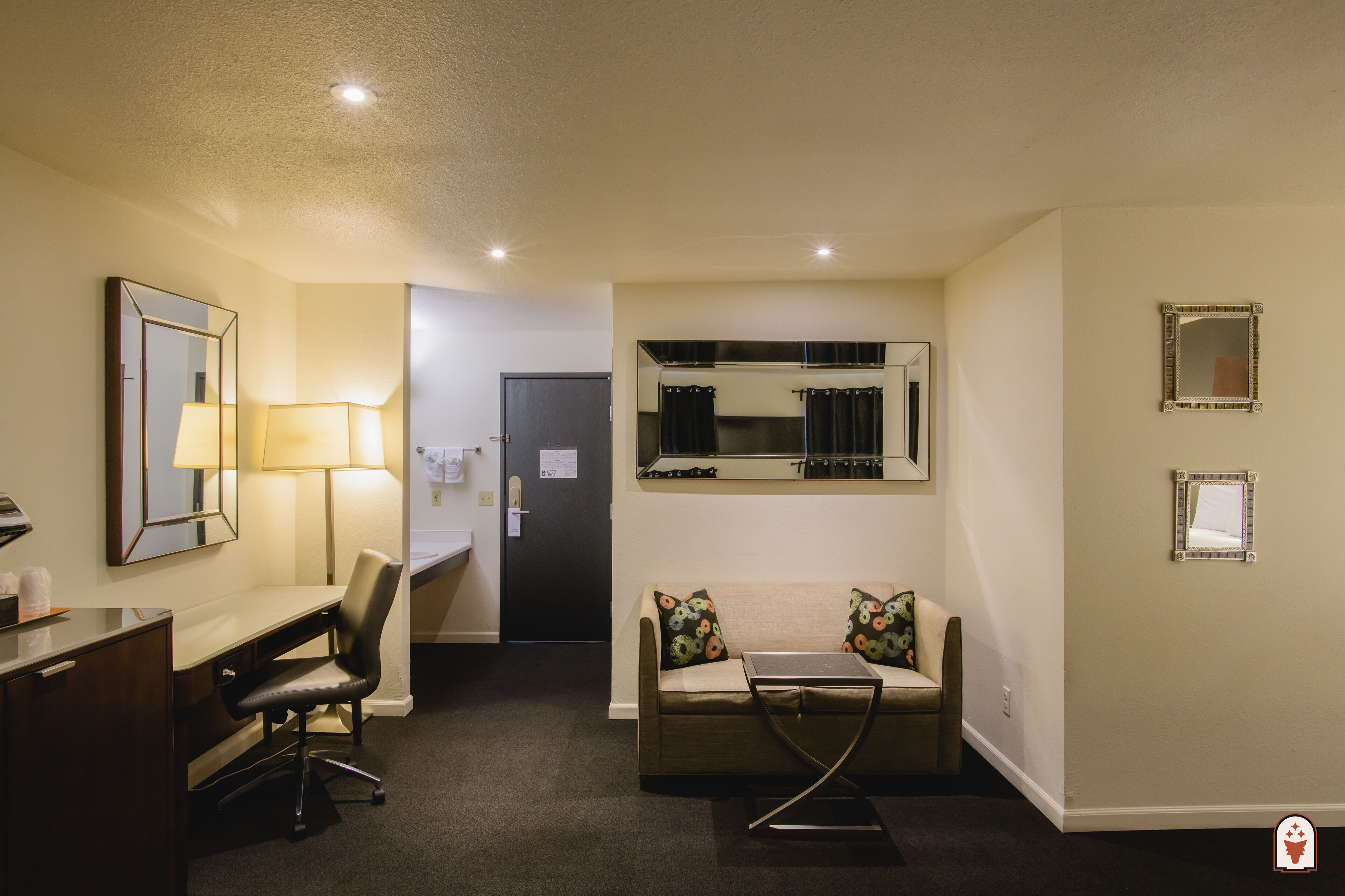 A hotel room with a desk, chair, sofa, and a small table. There are two mirrors on the walls and a door leading to another room.