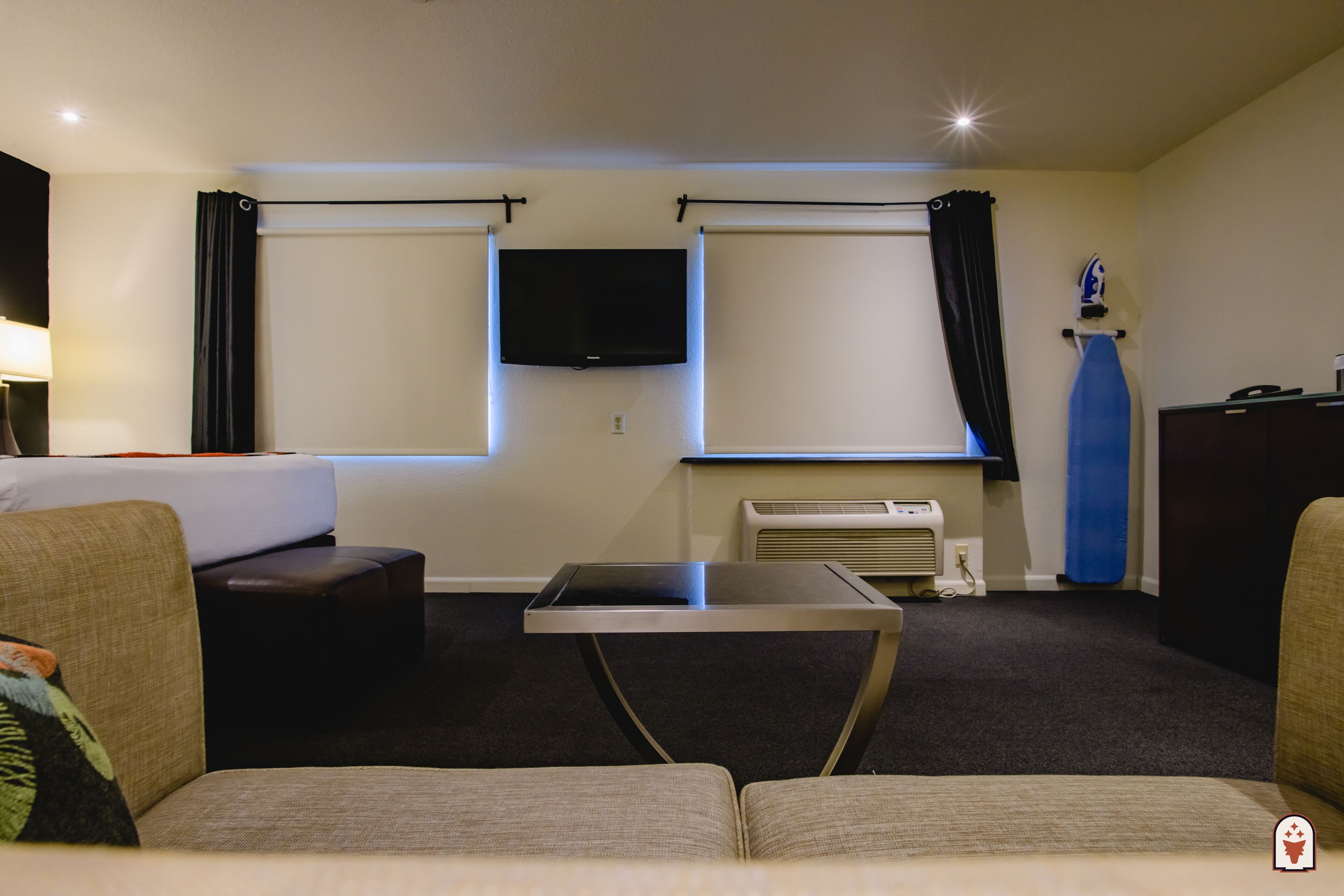 A hotel room with a sofa, a small table, a flat-screen TV mounted on the wall, and a window with curtains.