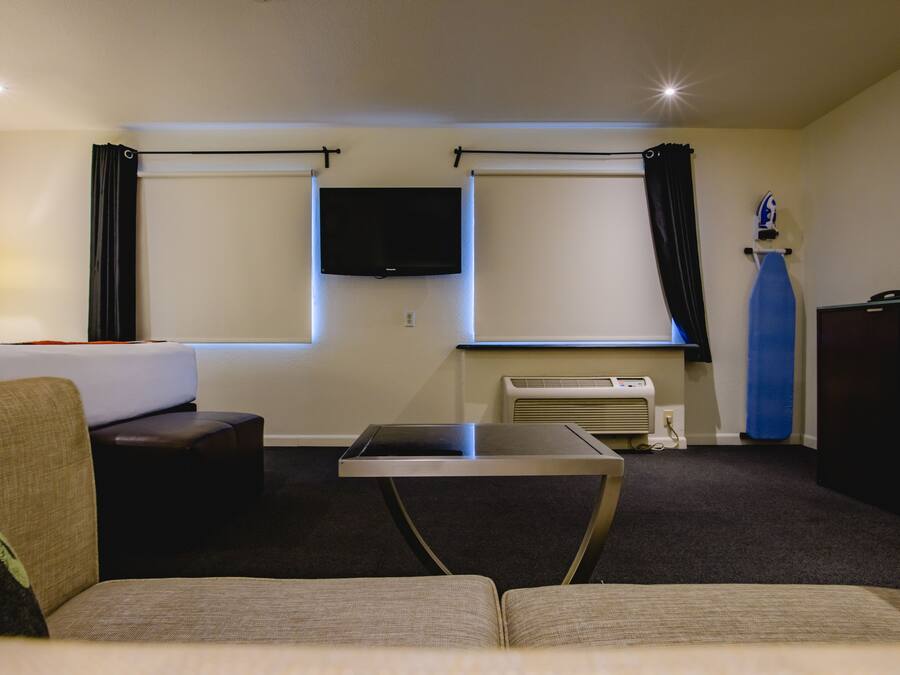 A hotel room with a sofa, a small table, a flat-screen TV mounted on the wall, and a window with curtains.