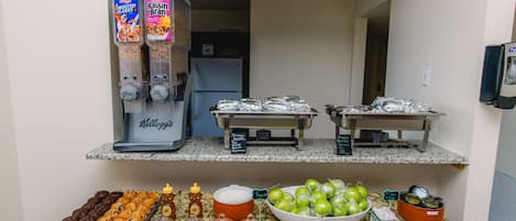 Uma bancada de cozinha com diversos alimentos e condimentos, incluindo uma cafeteira, uma tigela de maçãs verdes e uma bandeja de muffins.