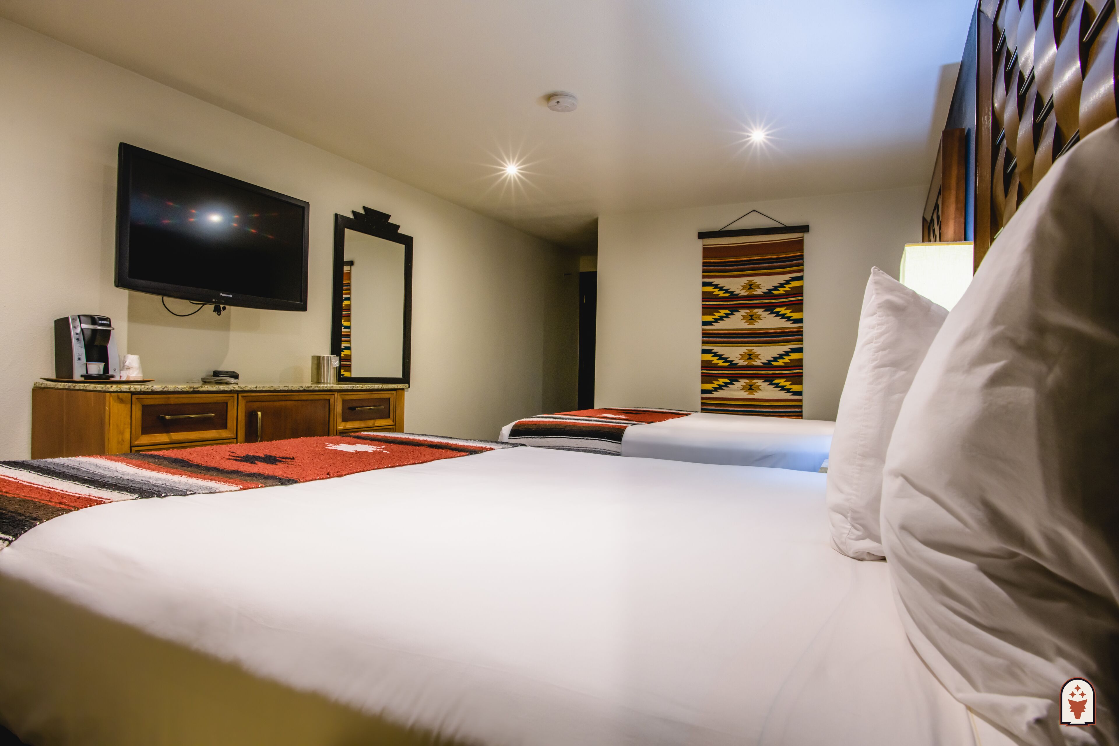 A hotel room with a large bed, a television, a coffee machine, and a patterned wall hanging.