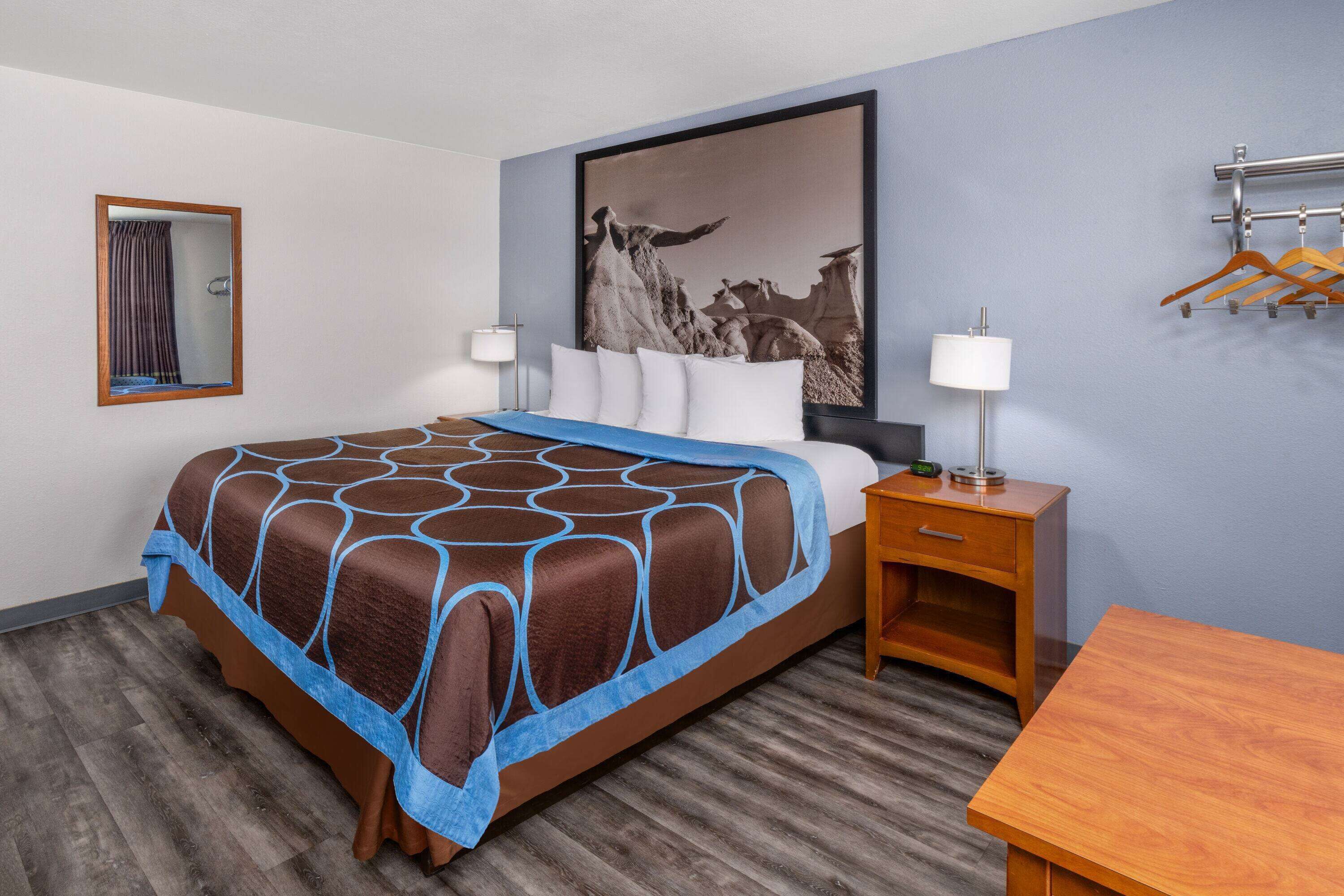Ein Hotelzimmer mit einem großen Bett, einem Nachttisch aus Holz, einem Schreibtisch, einem Kleiderhaken und einem gerahmten Bild an der Wand.