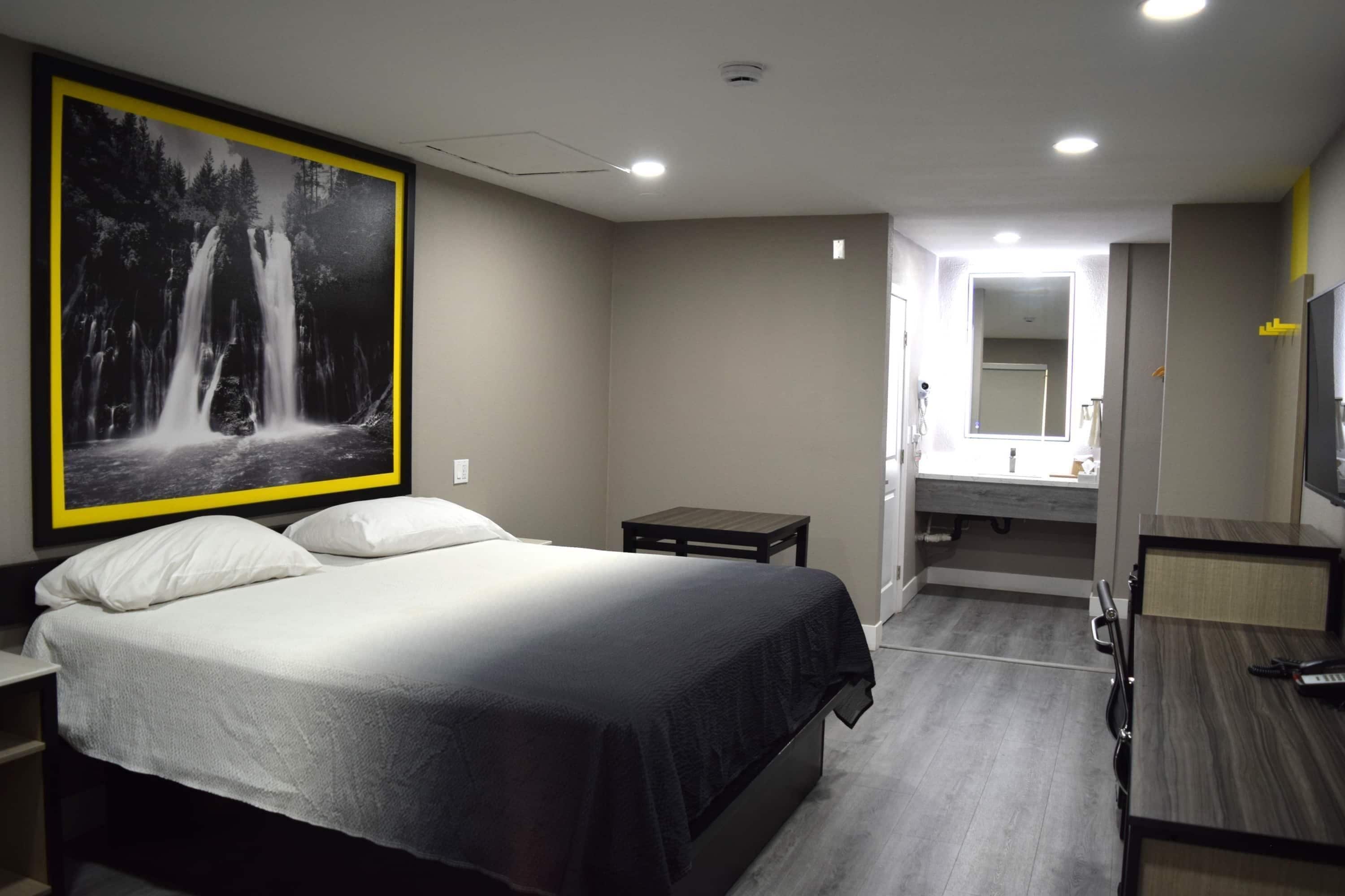 A hotel room with a large bed, a small bedside table, a mirror, a TV, and a bathroom with a sink and mirror.
