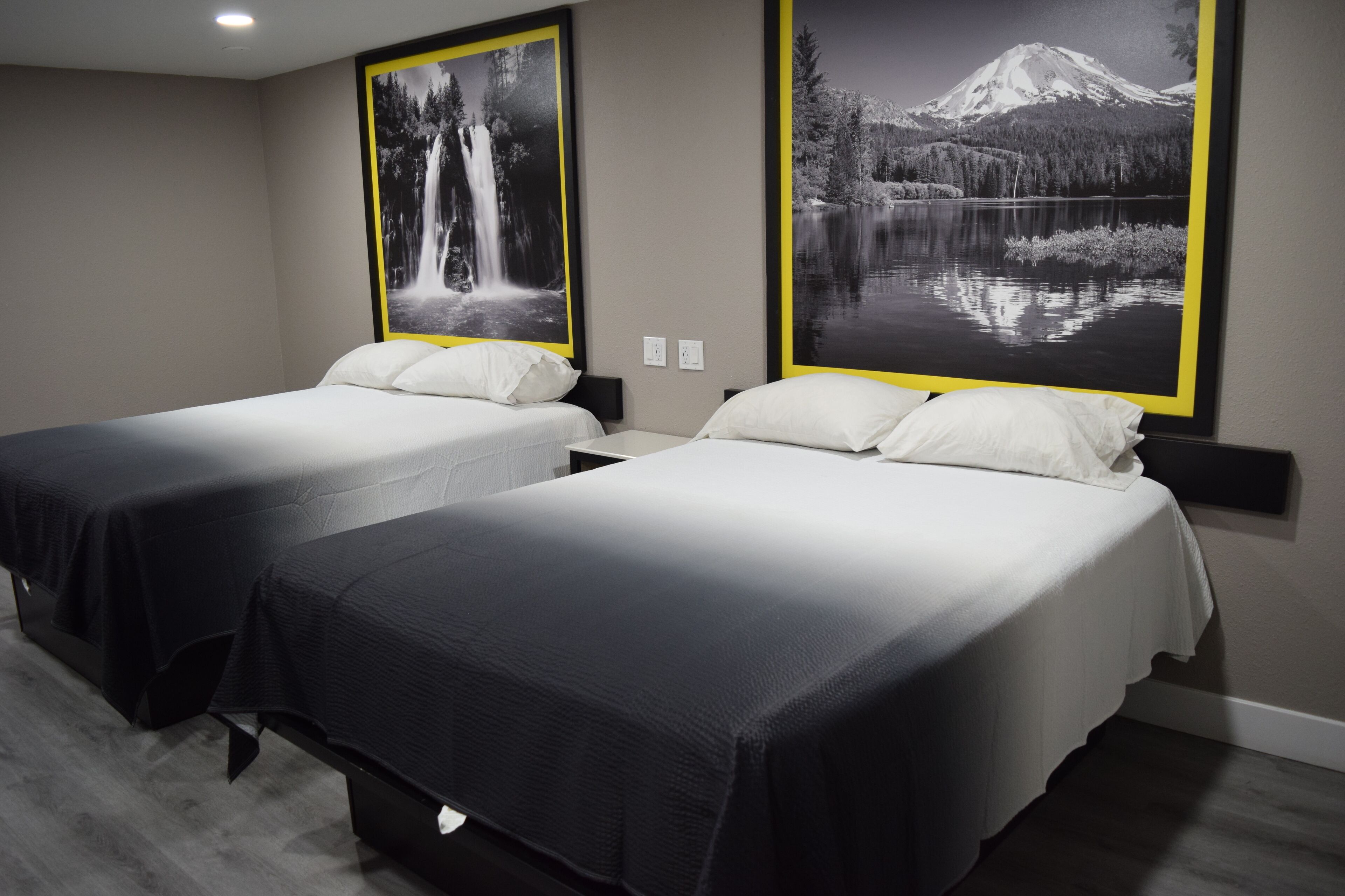 A hotel room with two beds, each with a black and white comforter, and large framed photographs of a waterfall and a mountain lake on the walls.