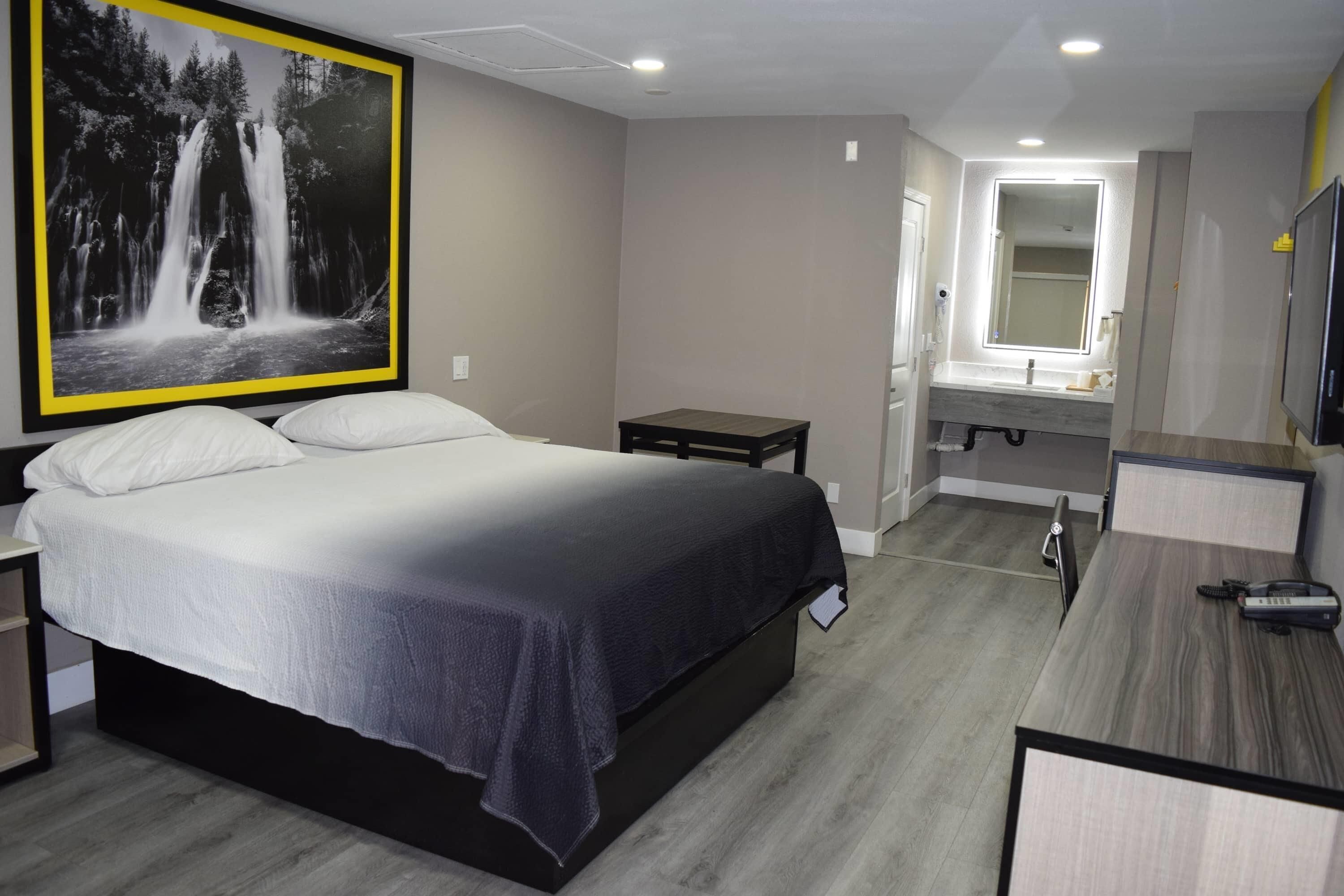 Een slaapkamer met een bed, nachtkastje, bureau, spiegel en een badkamer door een geopende deur zichtbaar.