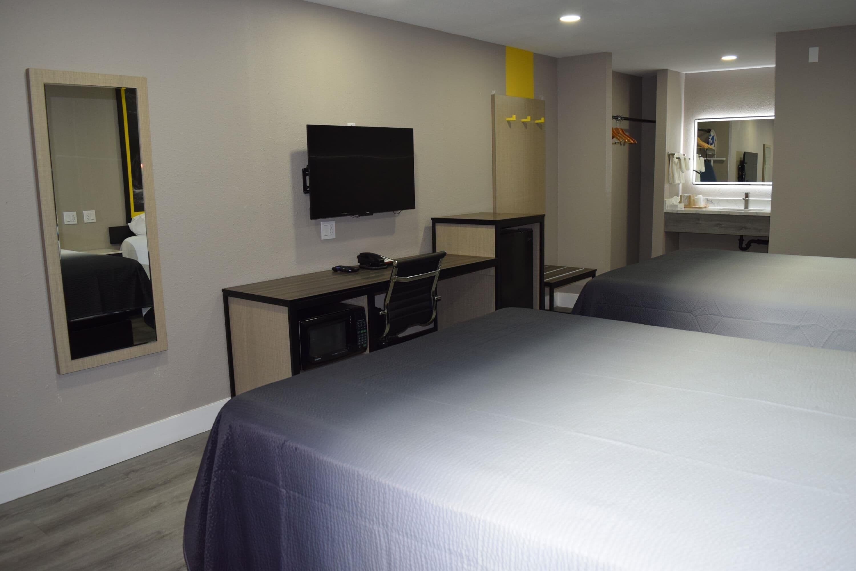 Quarto de hotel com duas camas, uma escrivaninha com cadeira, televisão, espelho e banheiro com pia e espelho.