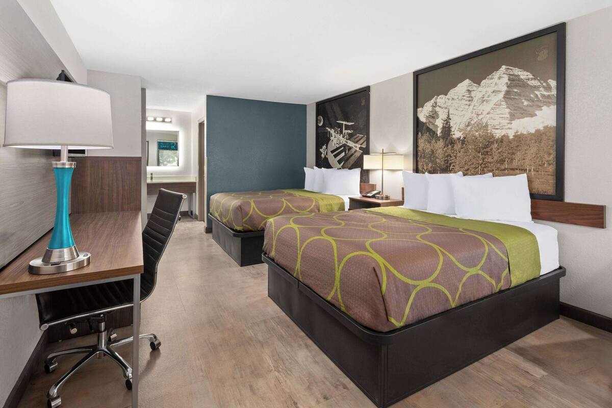 A hotel room with two beds, a desk, a chair, and a lamp.