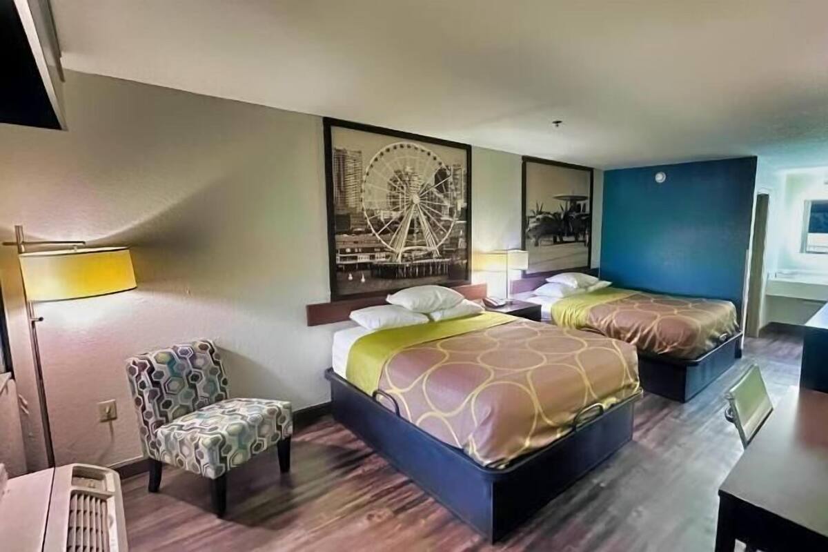 A hotel room with two beds, a chair, a desk, a lamp, and a wall with a large picture.