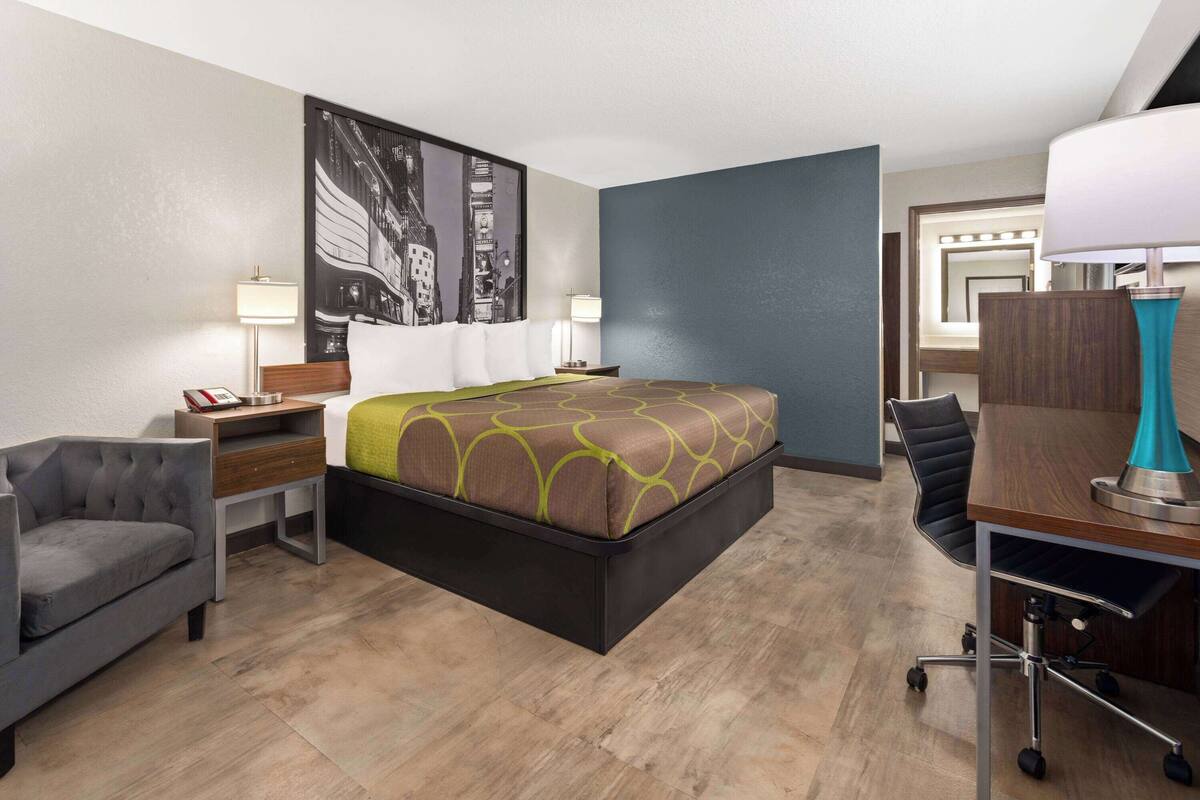 A modern hotel room with a large bed, a desk with a lamp, a chair, and a small table with a phone.